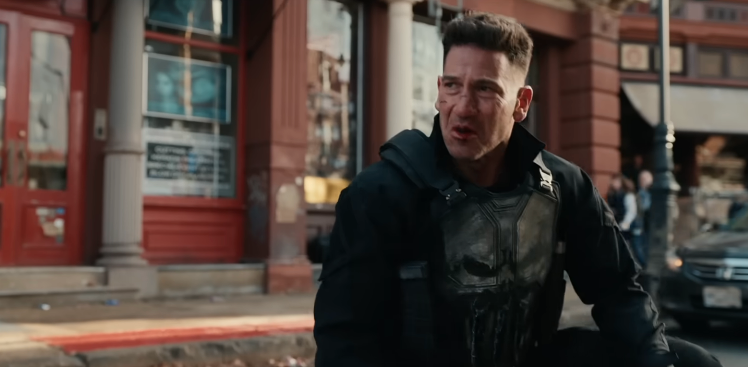 En mann med et bestemt uttrykk, iført taktisk utstyr med hodeskalleemblem, befinner seg i en gate.