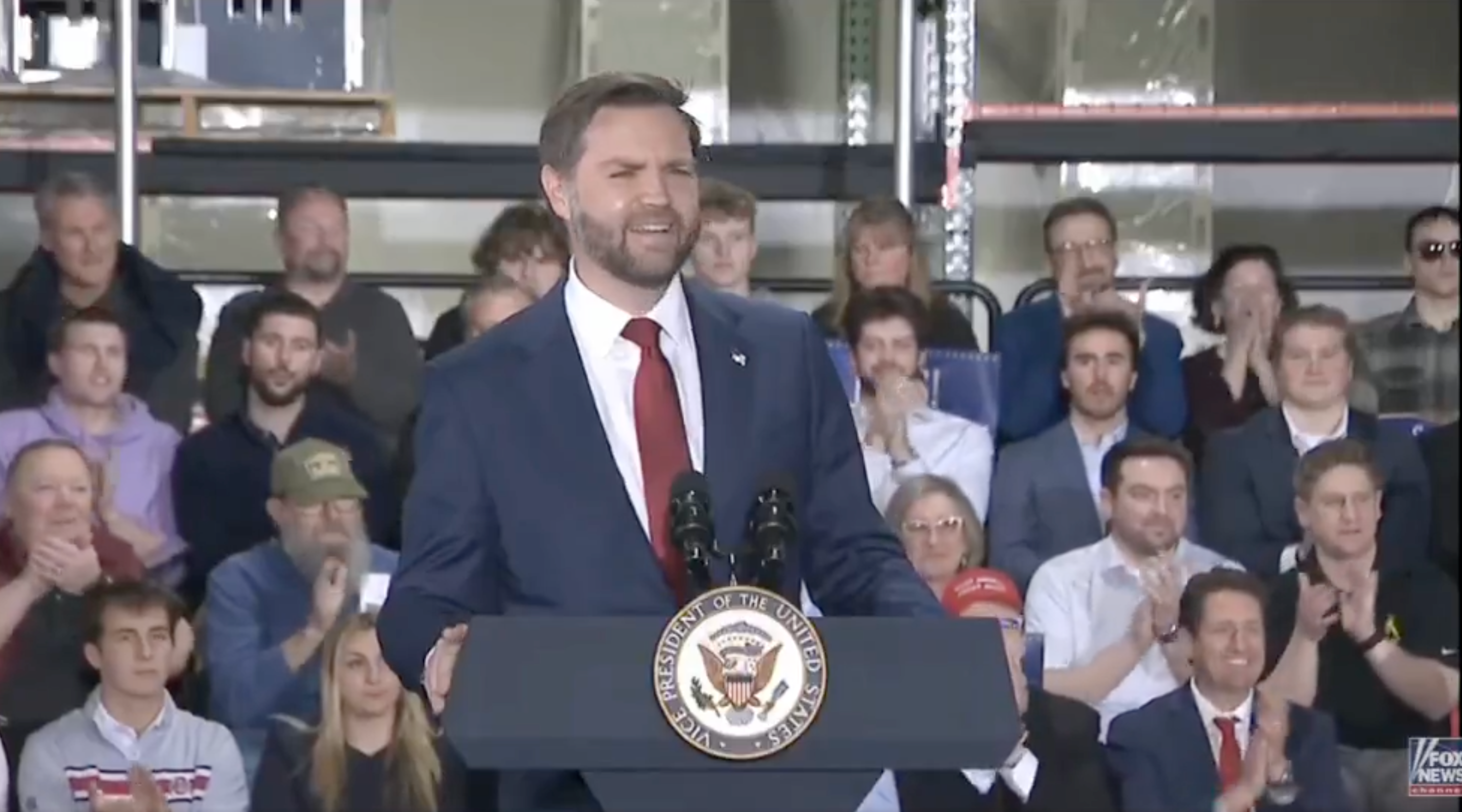 A antheral   successful  a suit   speaks astatine  a podium with a statesmanlike  seal, addressing an assemblage  sitting and applauding down  him