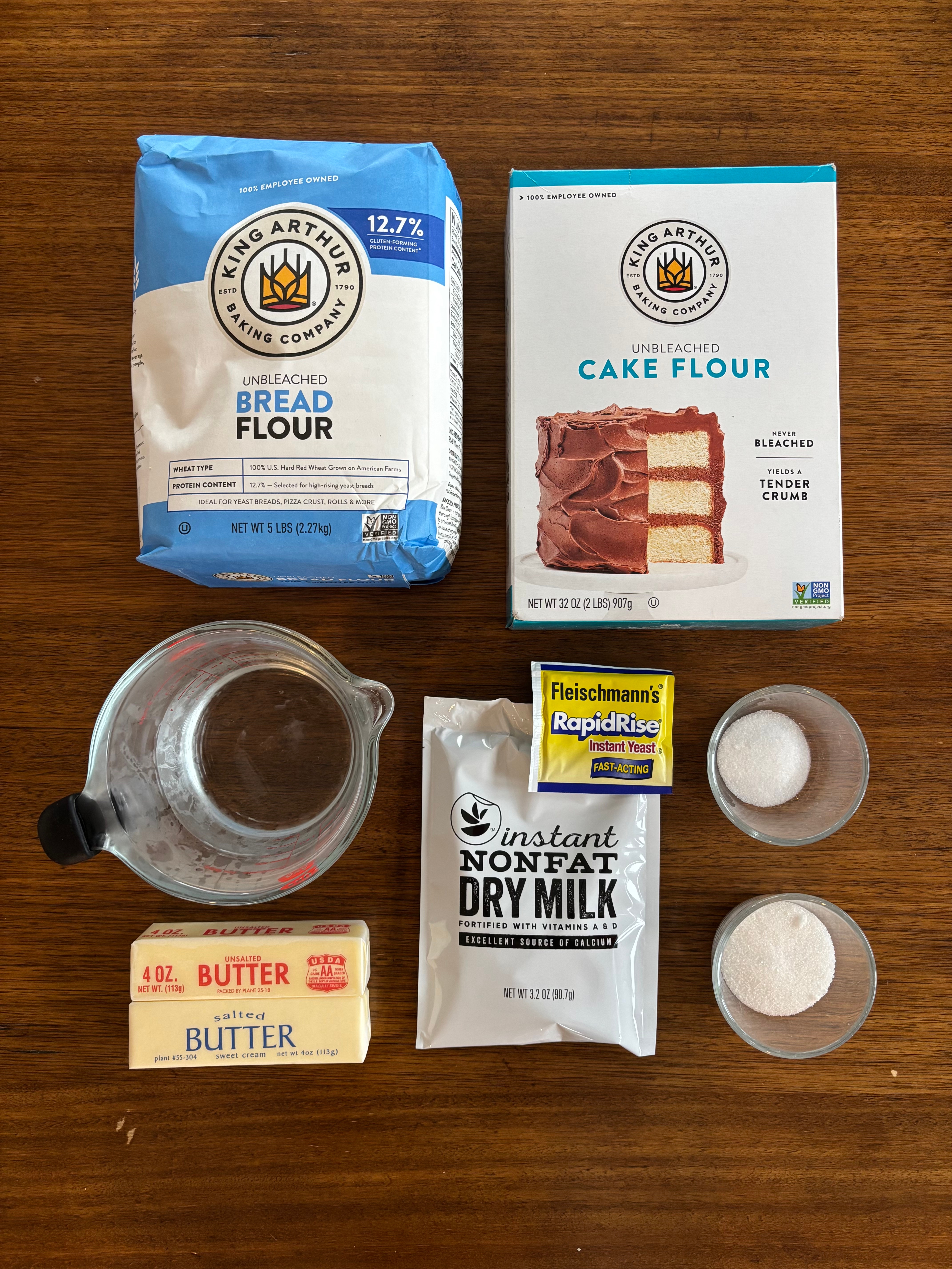 Various baking ingredients connected  a woody  table, including breadstuff  flour, barroom   flour, instant adust  milk, butter, yeast, and tiny  bowls of sweetener  and salt