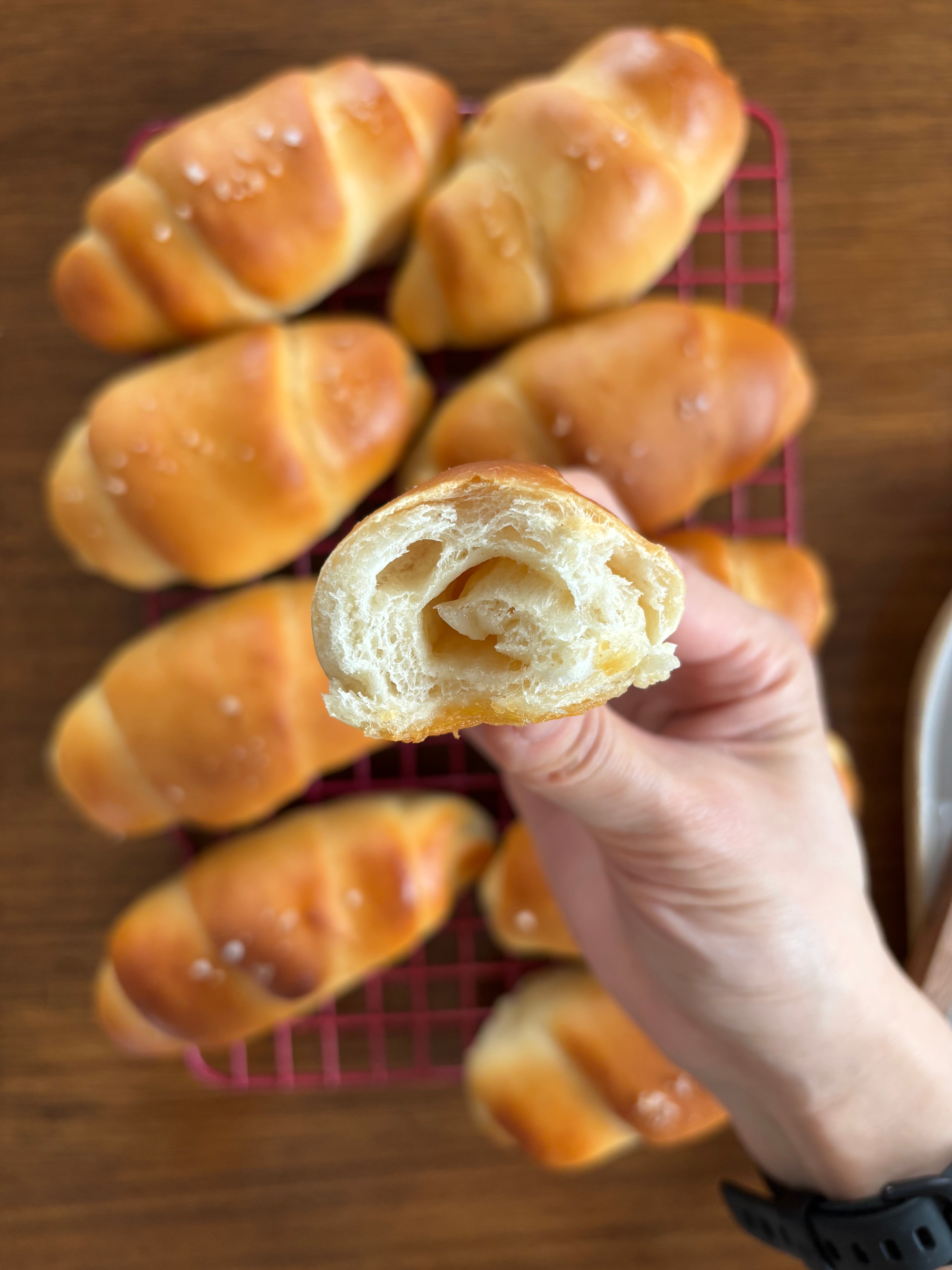 Close-up of a manus  holding a flaky freshly baked croissant, with much  croissants cooling connected  a rack successful  the background