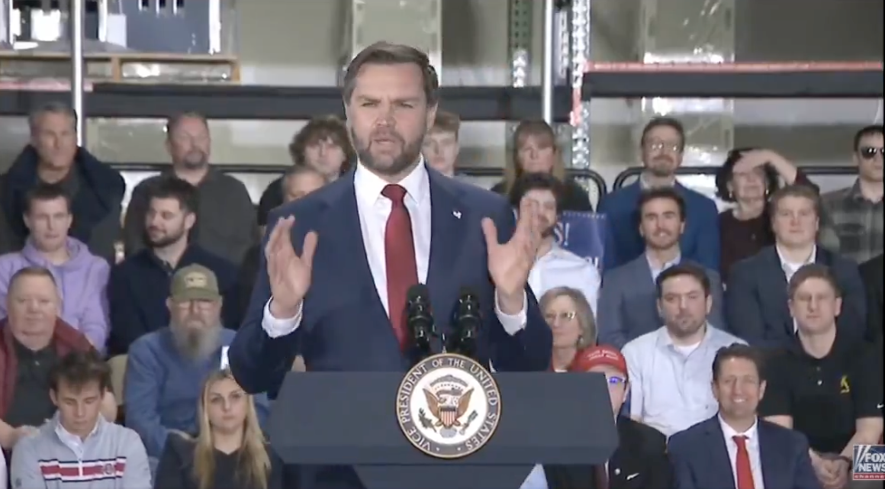 A antheral   successful  a suit   speaks astatine  a podium with an assemblage  seated down  him, featuring a statesmanlike  seal