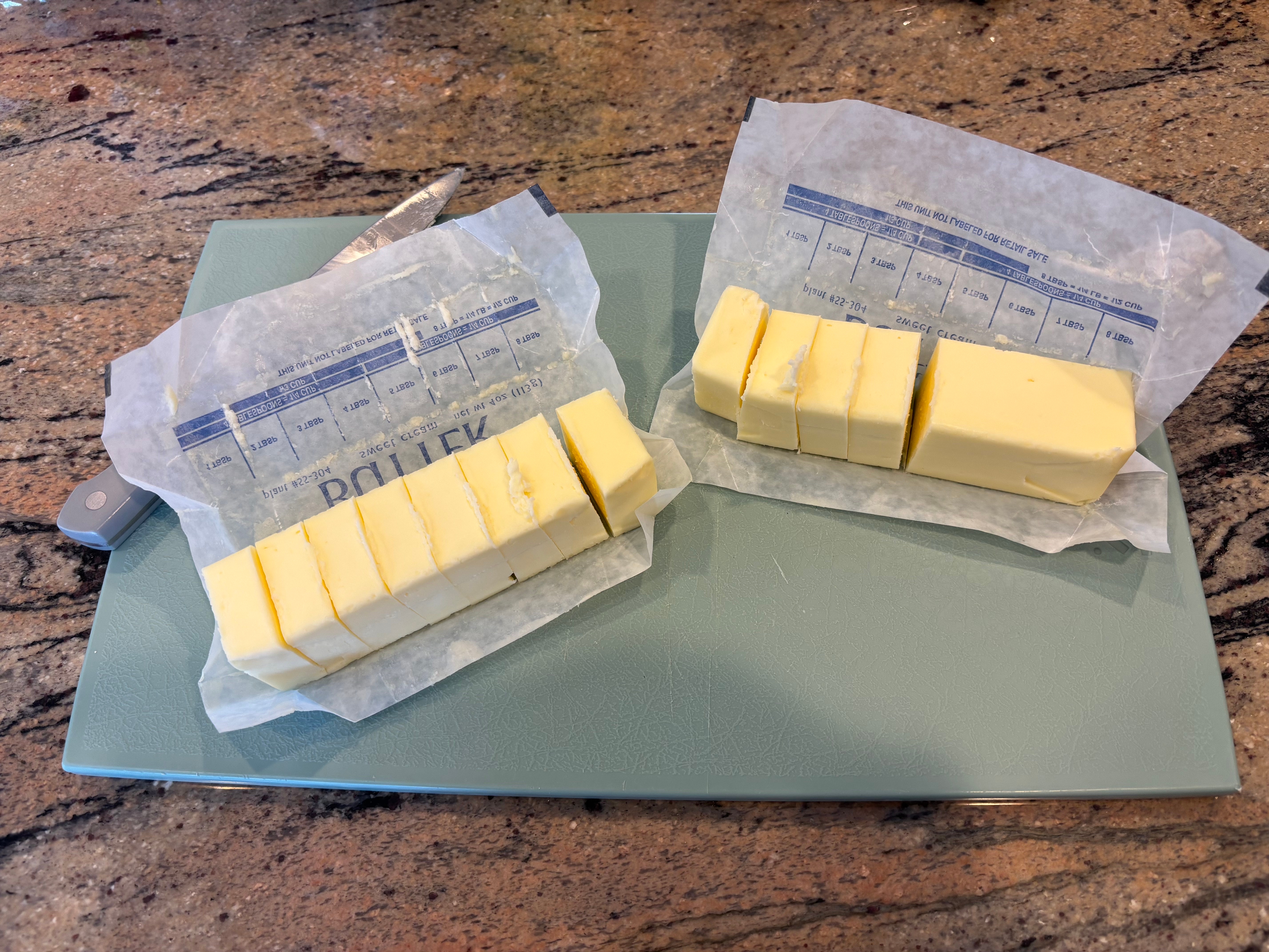 Two sticks of food  partially sliced connected  opened wrappers, resting connected  a cutting board