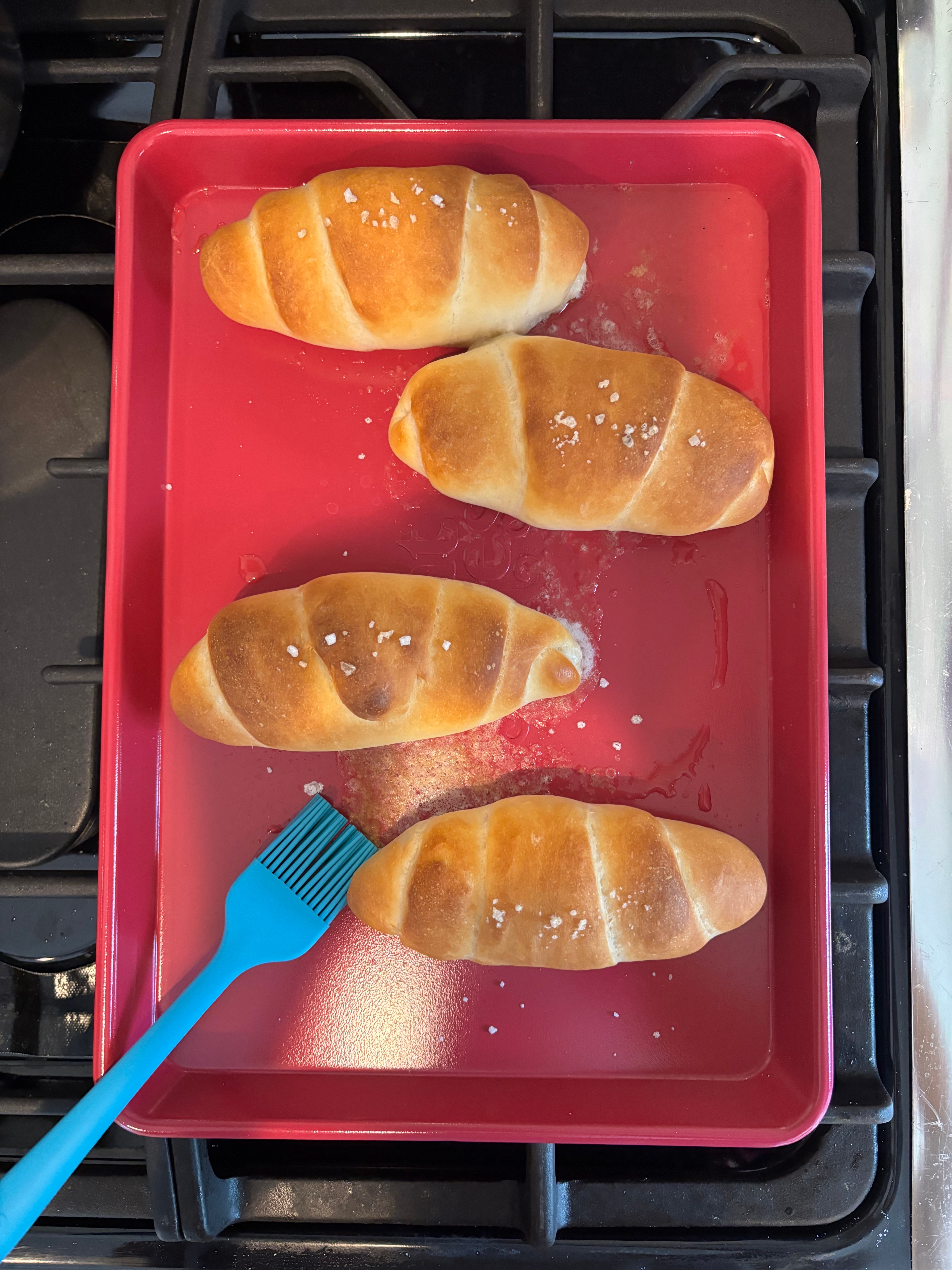 Four baked crescent rolls connected  a tray, sprinkled with salt, alongside a bluish  brush