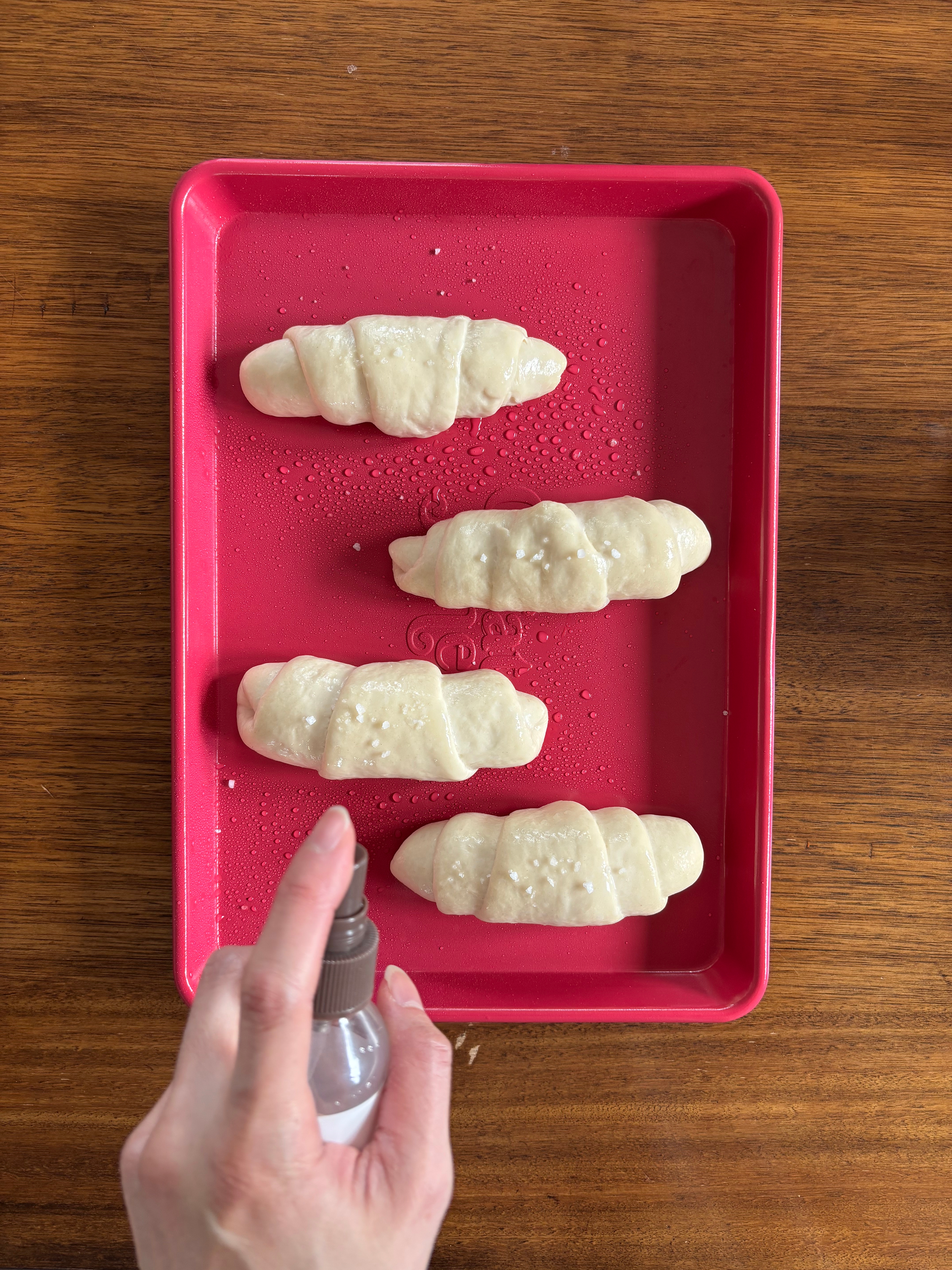Four uncooked croissants connected  a tray being sprayed with water