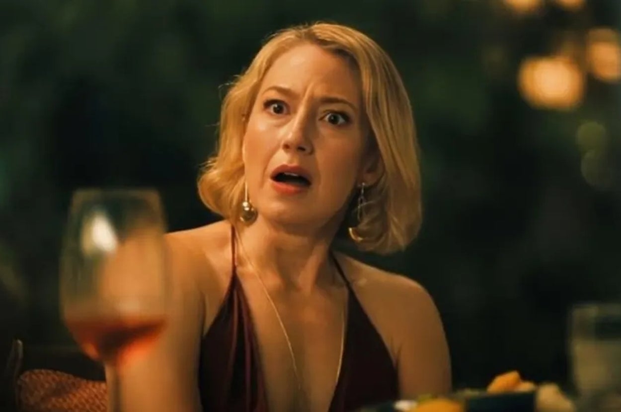 Person at a dinner table looks surprised, sitting outdoors with evening lighting. Elegant outfit and jewelry suggest a social or formal gathering