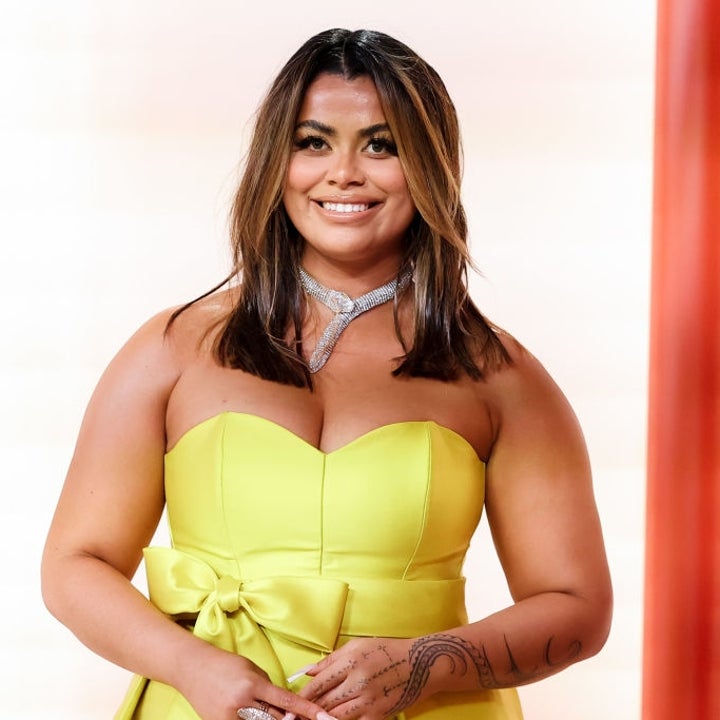 Person in a strapless gown with a bow and necklace on a red carpet, smiling at the camera