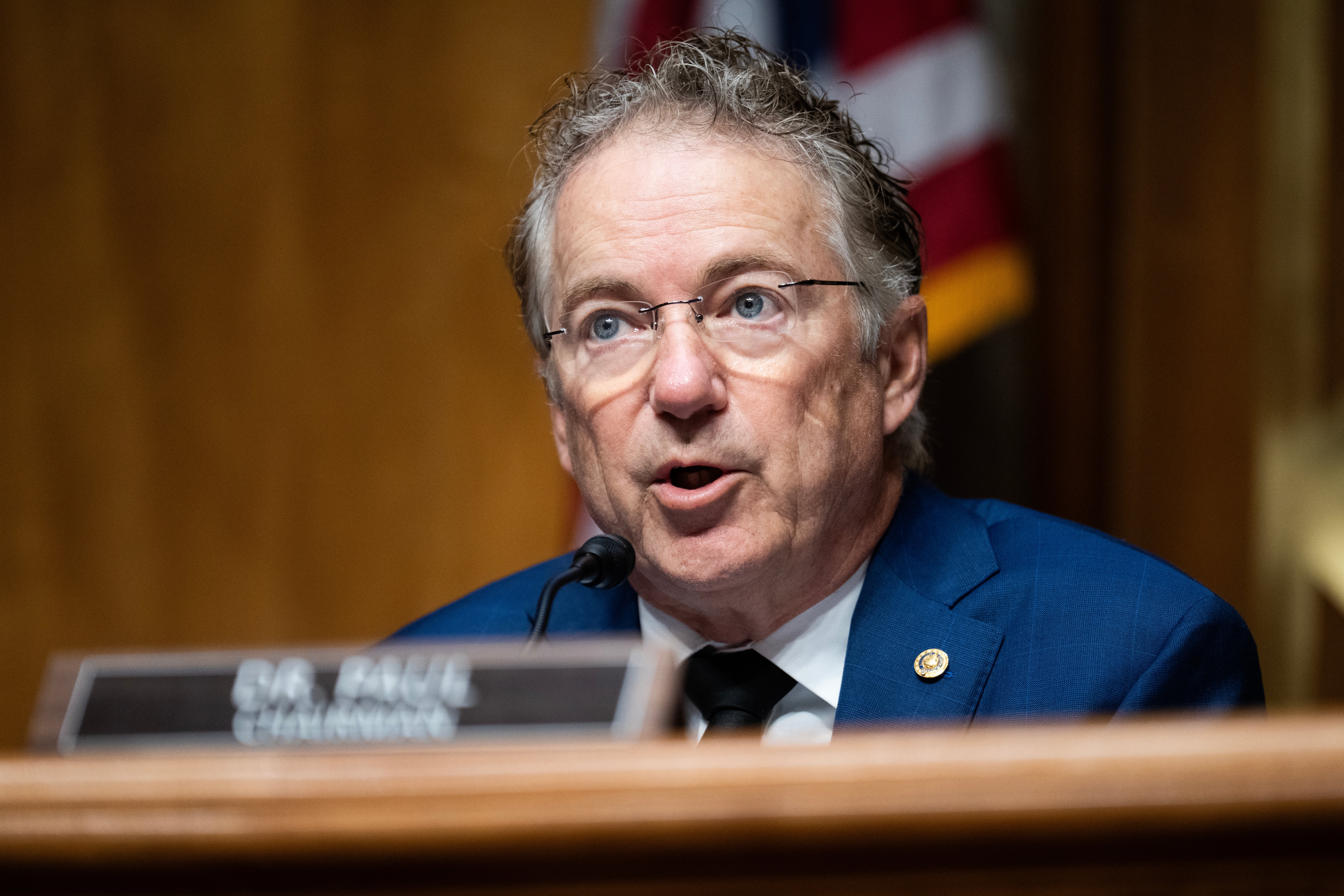 En mann i blå dress snakker i en mikrofon ved en formell begivenhet. Navneskiltet hans lyder 