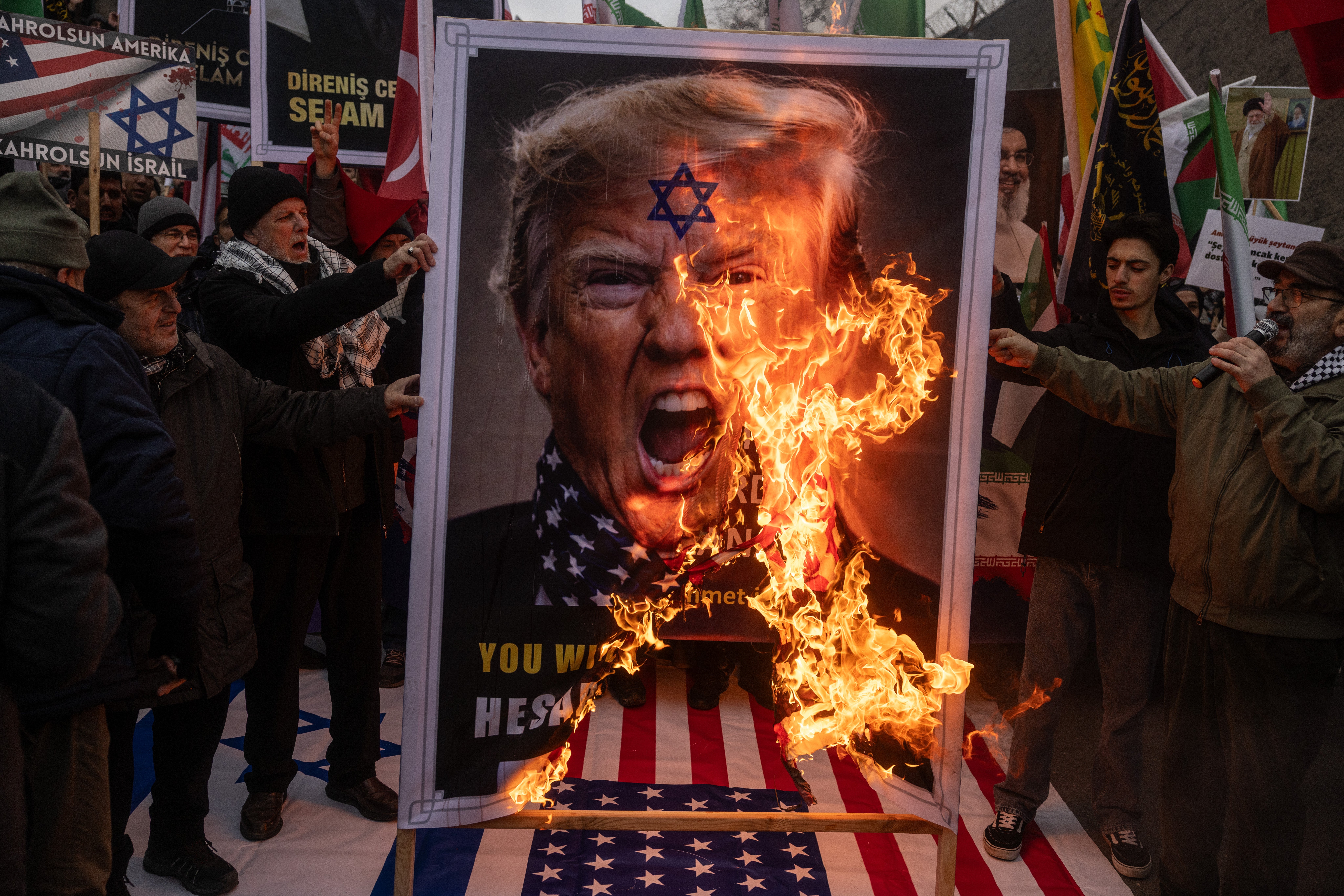 Demonstranter brenner en stor plakat som portretterer en politisk skikkelse med tilleggssymboler, stående på et amerikansk flagg under en demonstrasjon