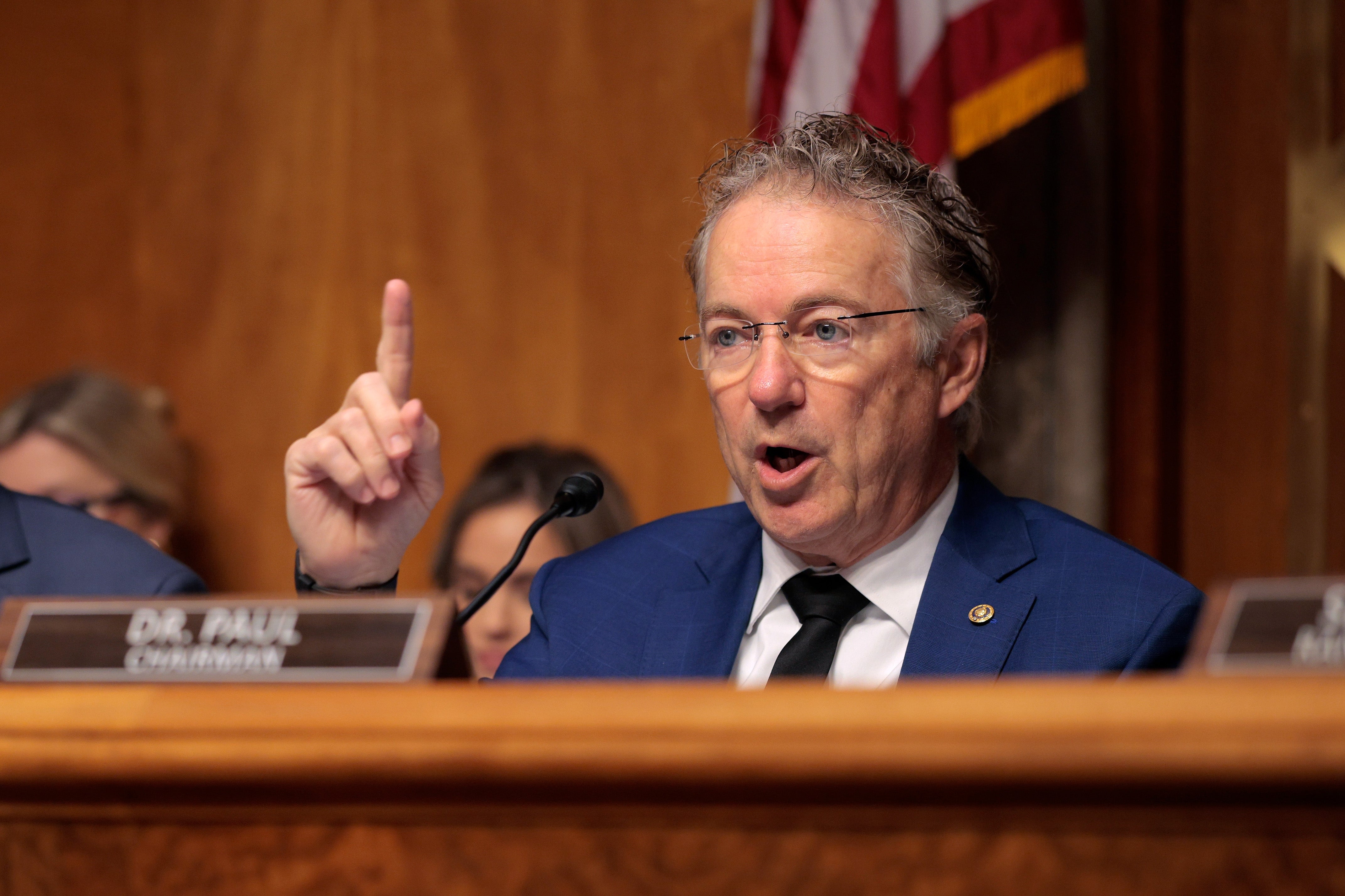 En passende mann snakker lidenskapelig under en regjeringshøring, gestikulerer med en løftet finger, sittende foran et navneskilt