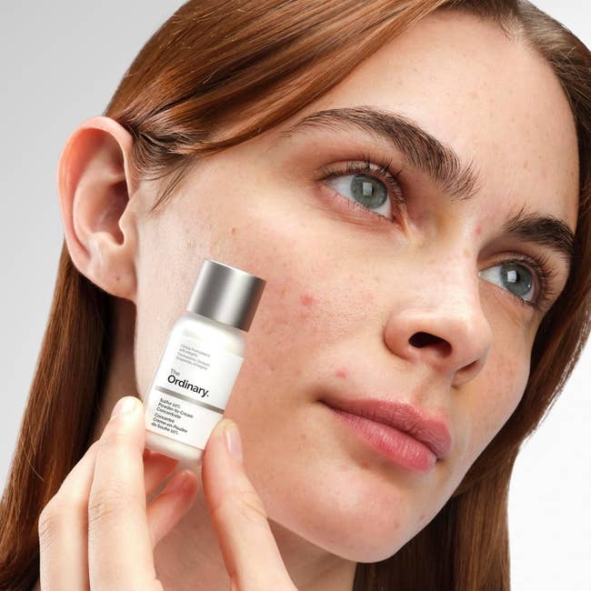 A idiosyncratic   with wide   tegument  and flimsy  blemishes holds a tiny  vessel  of The Ordinary skincare merchandise  adjacent   their face