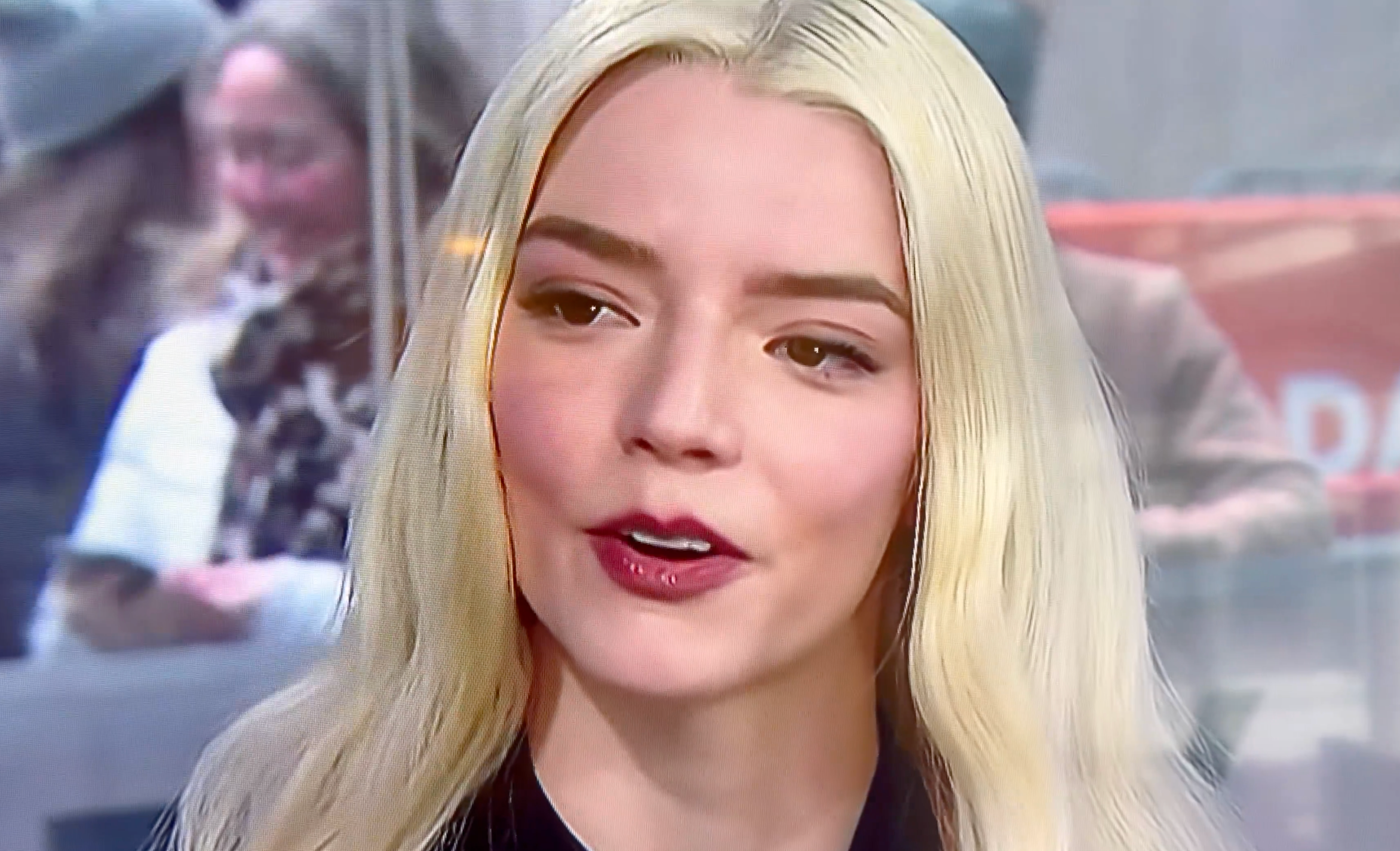 A idiosyncratic   with long, light-colored hairsbreadth  is talking. Background shows blurred radical   and outdoor setting