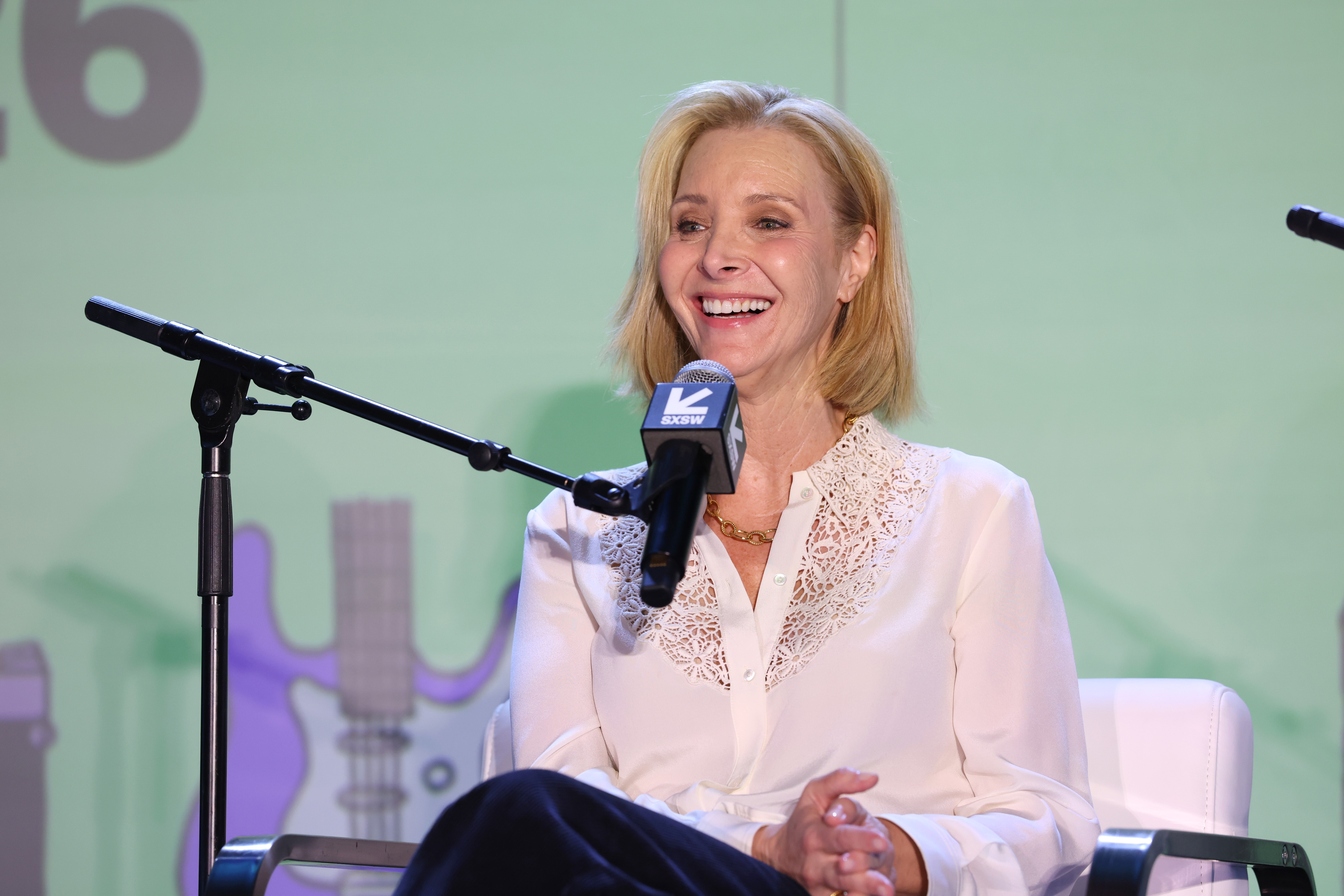 A idiosyncratic   sits and smiles during a sheet  discussion, wearing a achromatic  lace-collared blouse and navy pants, talking into a microphone