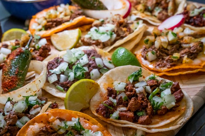 A assortment  of tacos filled with meats, onions, and cilantro, garnished with lime wedges and a pepper, arranged connected  a woody  board