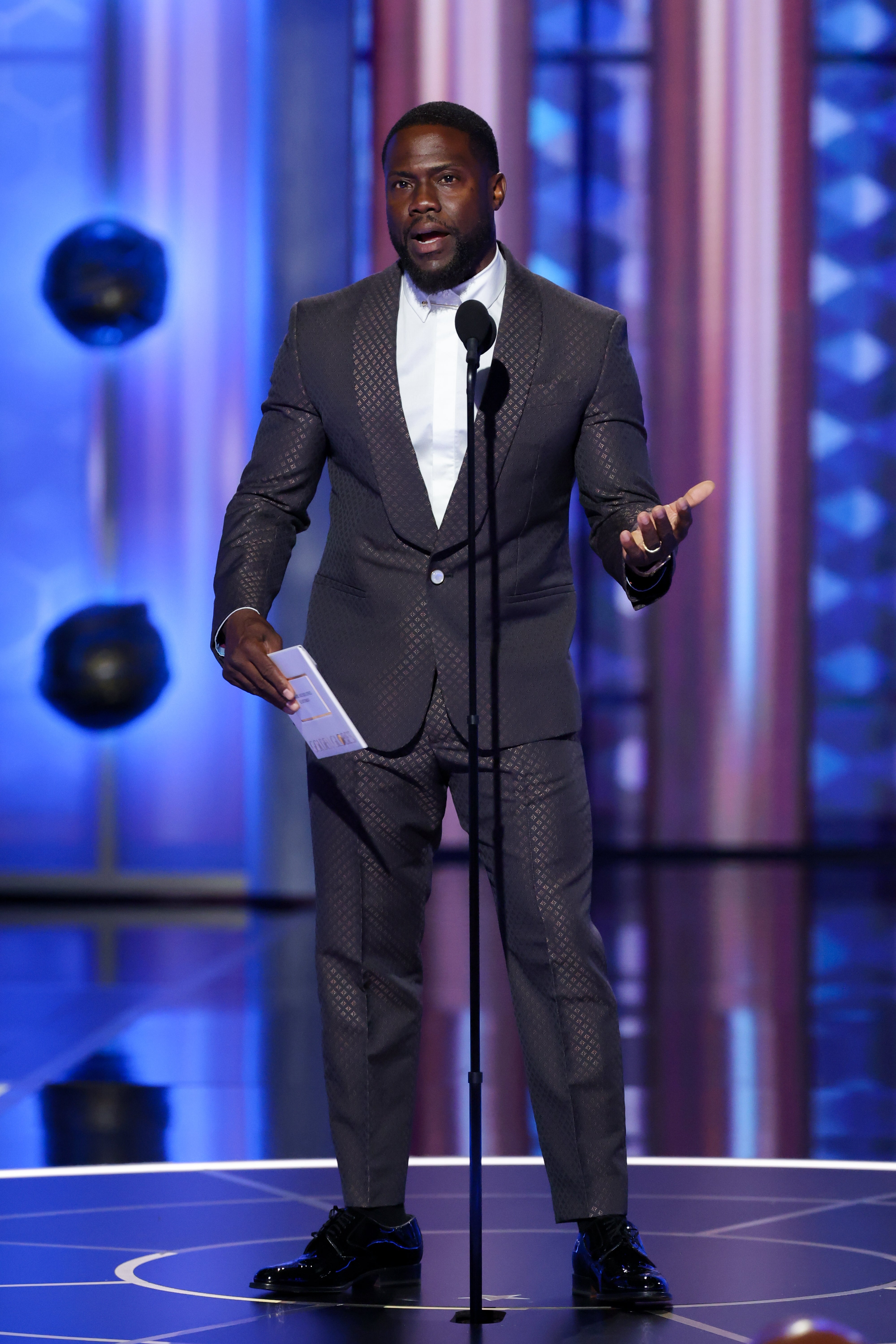 En person i en mønstret dress snakker på scenen, gestikulerer med den ene hånden, holder et kort med den andre, under en formell begivenhet