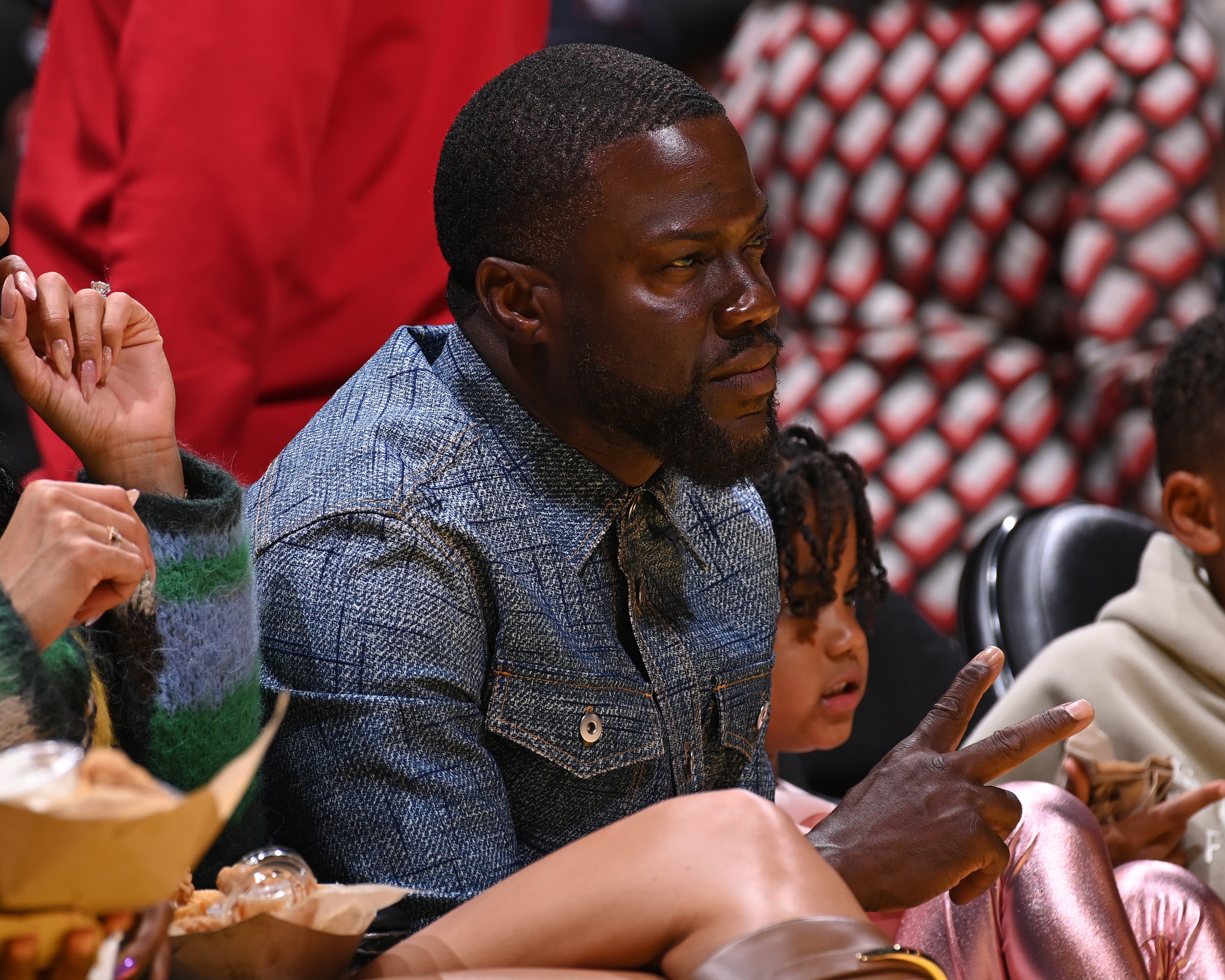 En person sitter på banen ved en basketballkamp, ​​iført en dongerijakke, og virker engasjert i spillet. Folk sitter i nærheten