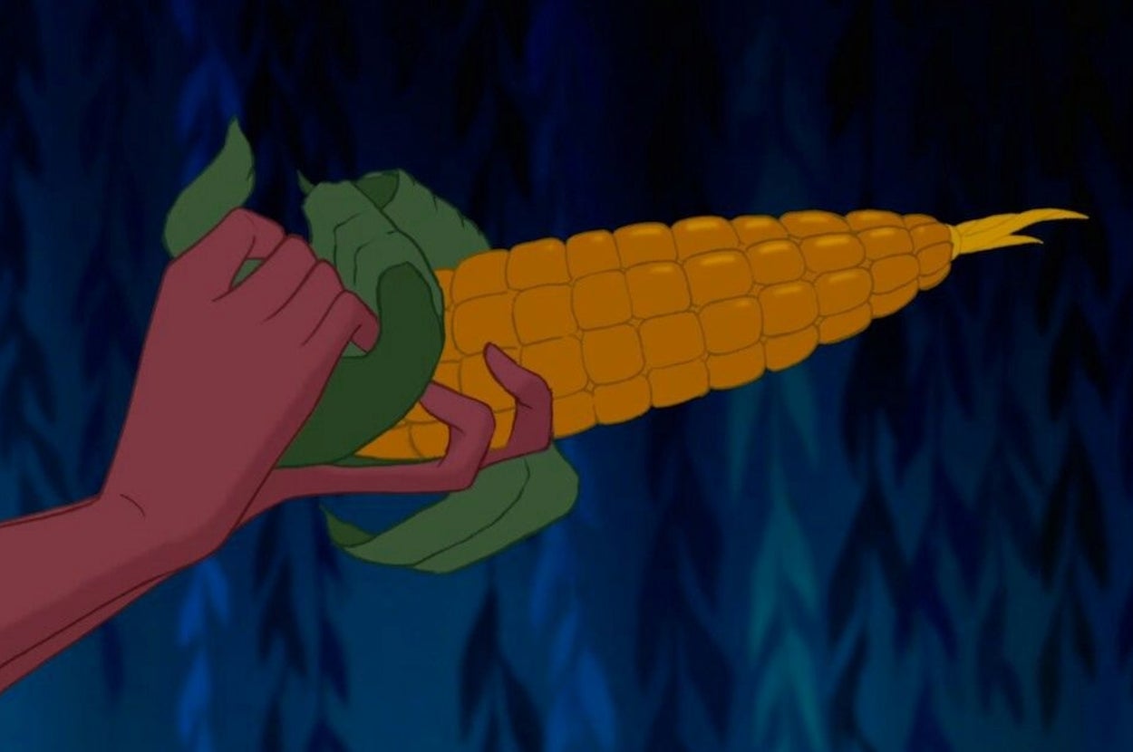 An animated hand holds an ear of corn partially wrapped in green husk against a dark background
