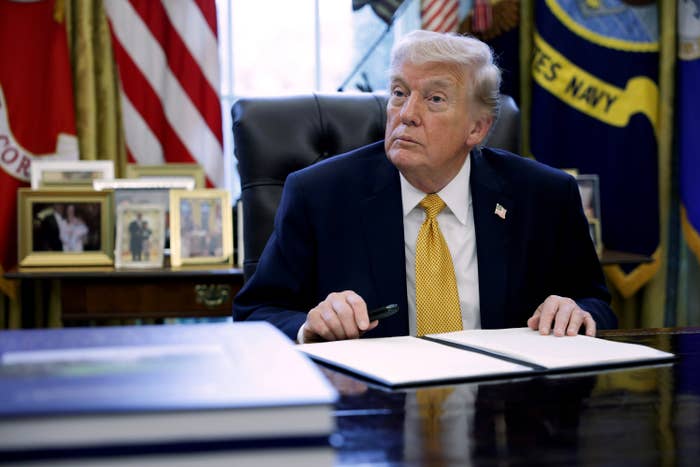 A idiosyncratic   successful  a suit   sits astatine  a table  successful  an authoritative  mounting  with flags and framed photos successful  the background, holding a pen implicit    a document