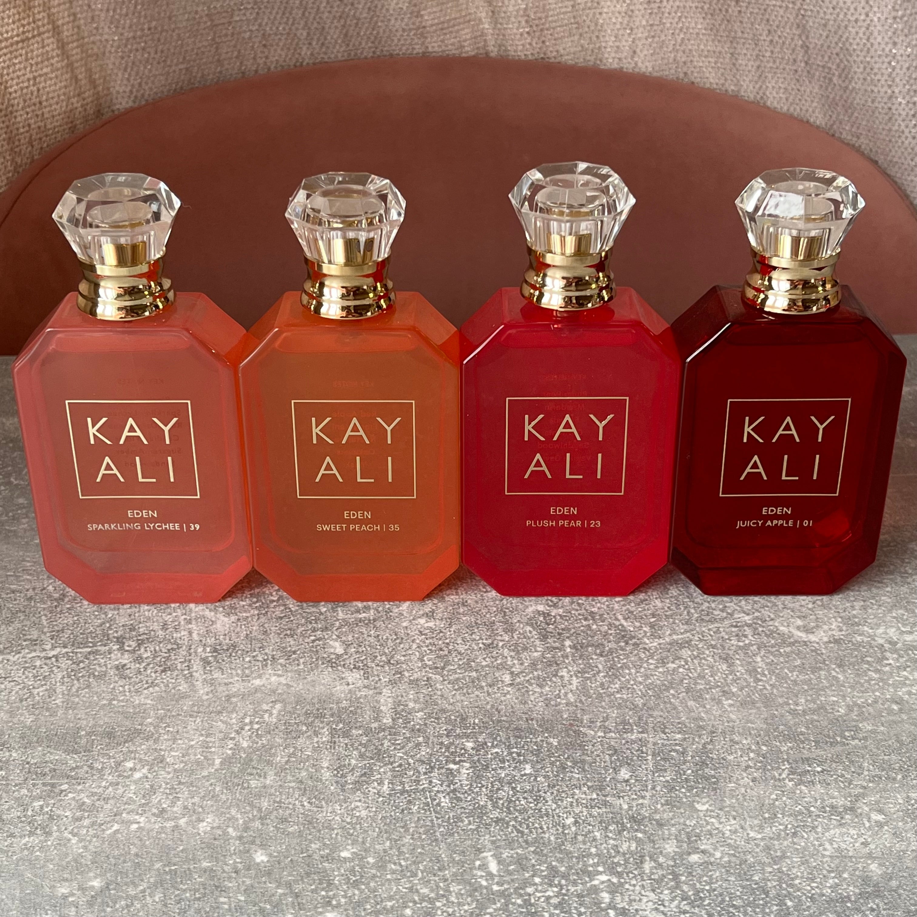 Four Kayali fragrance bottles aligned on a textured surface: Sparkling Lychee, Sweet Peach, Blush Peony, and Juicy Apple
