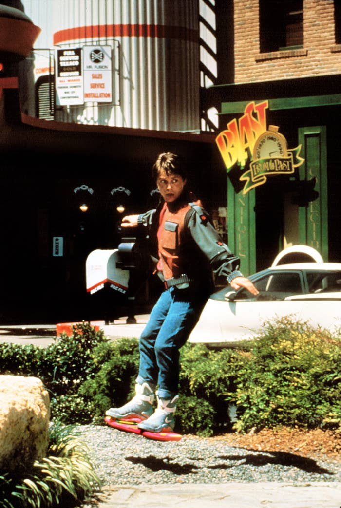 A idiosyncratic   rides a hoverboard connected  a street, wearing a futuristic outfit and looking surprised. The country   is from a fashionable  '80s sci-fi movie