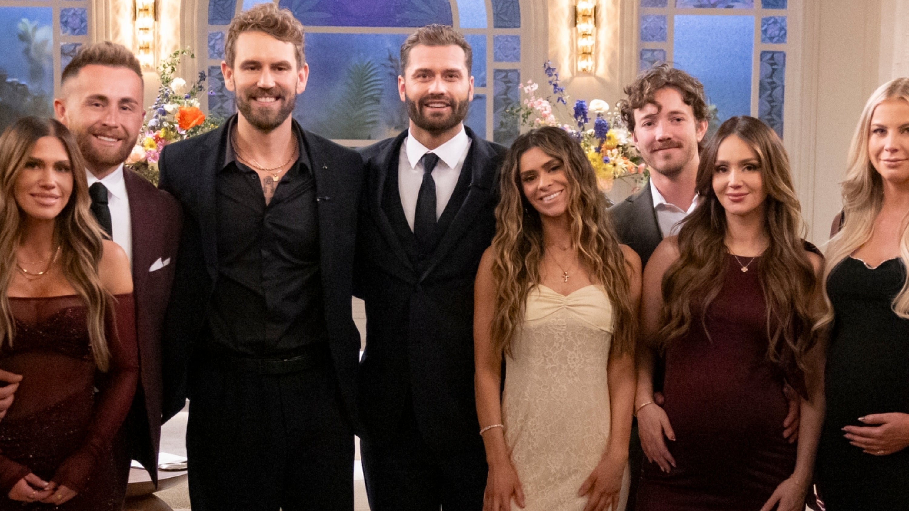 En gruppe mennesker i kveldstøy står i et godt opplyst rom med buede vinduer. Noen individer har synlige babyhumper, noe som tyder på graviditet