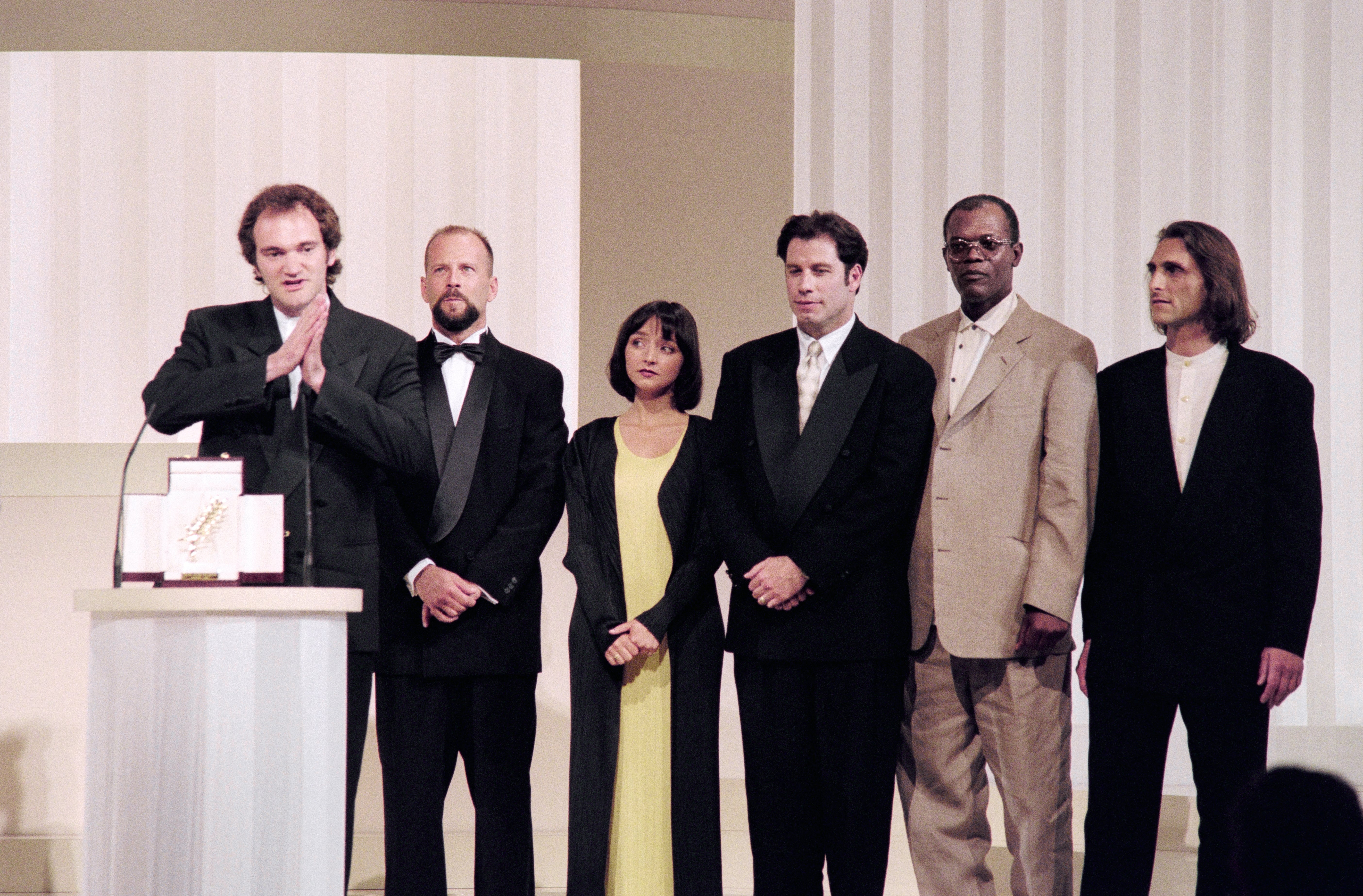 Quentin Tarantino and a Group of six people on stage wearing formal attire, with the man at a podium speaking, possibly at an award ceremony
