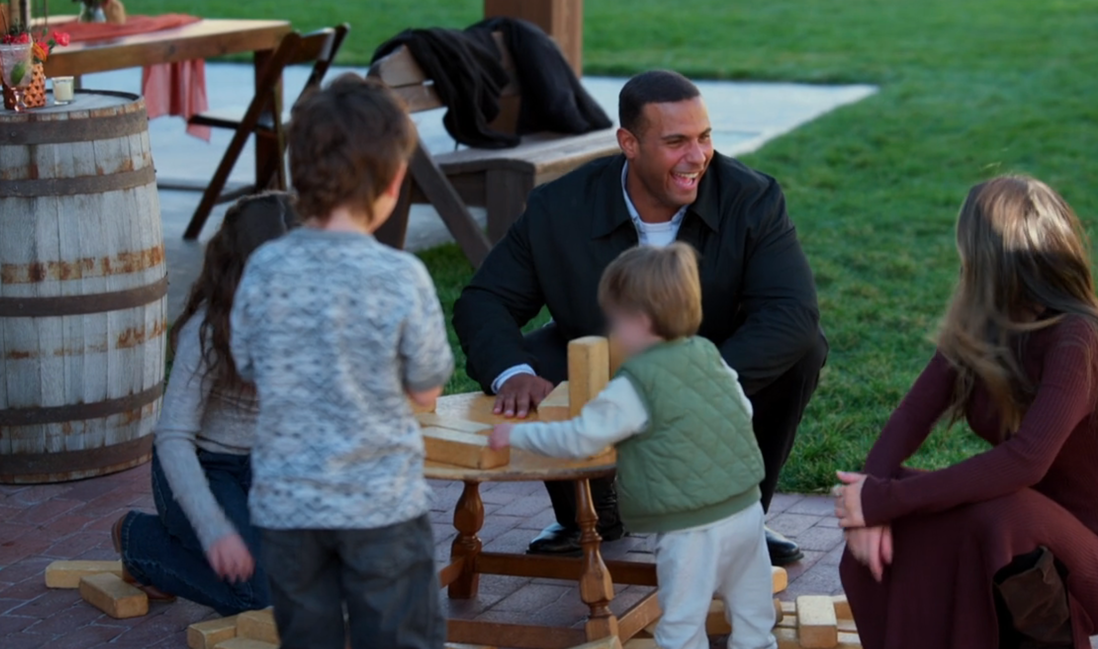 En gruppe voksne og barn leker med treklosser utendørs. En mann huker seg smilende mens en kvinne og to barn engasjerer seg i spillet