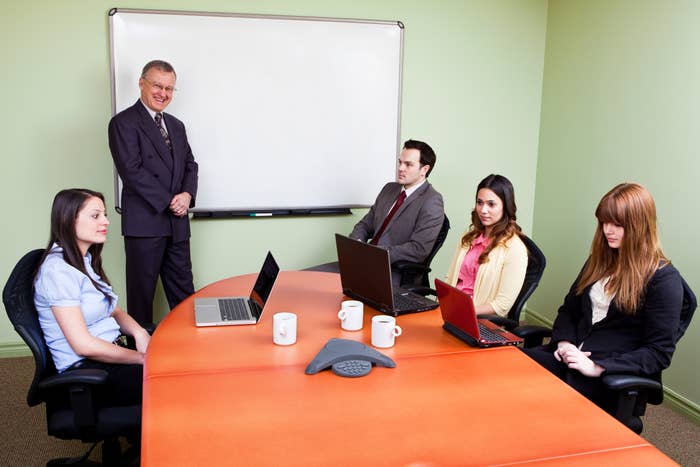 Five radical   successful  a league  room, 3  seated astatine  a array  with laptops, 1  lasting  by a whiteboard, and 1  sitting with hands connected  lap