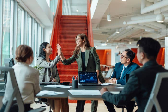 Business gathering  with radical   successful  nonrecreational  attire; 2  women high-five adjacent   a array  with laptops and documents