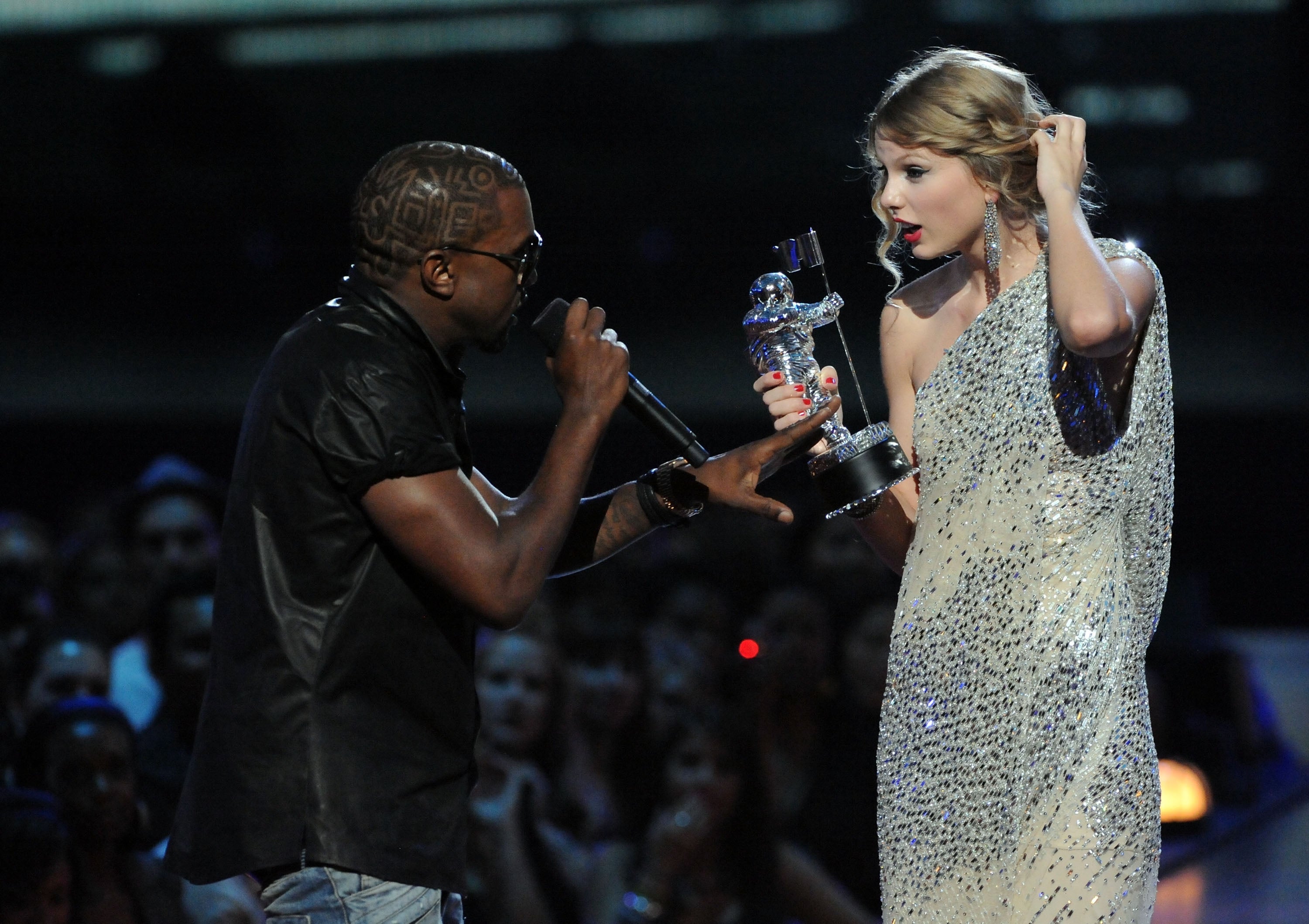 Kanye West interrupts an award acceptance on stage. Another stands holding a trophy, looking surprised. Crowd visible in background