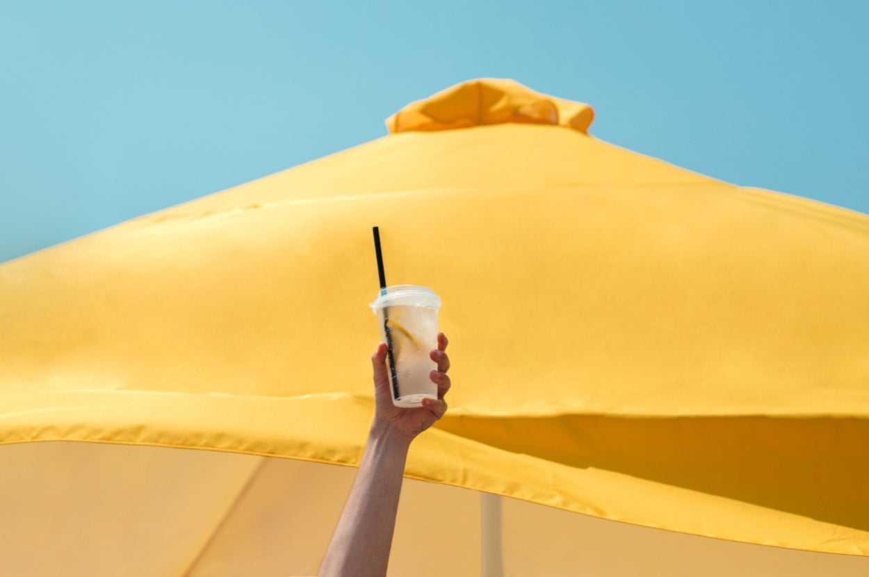 A hand holds a plastic cup with a straw against a backdrop of a large outdoor umbrella