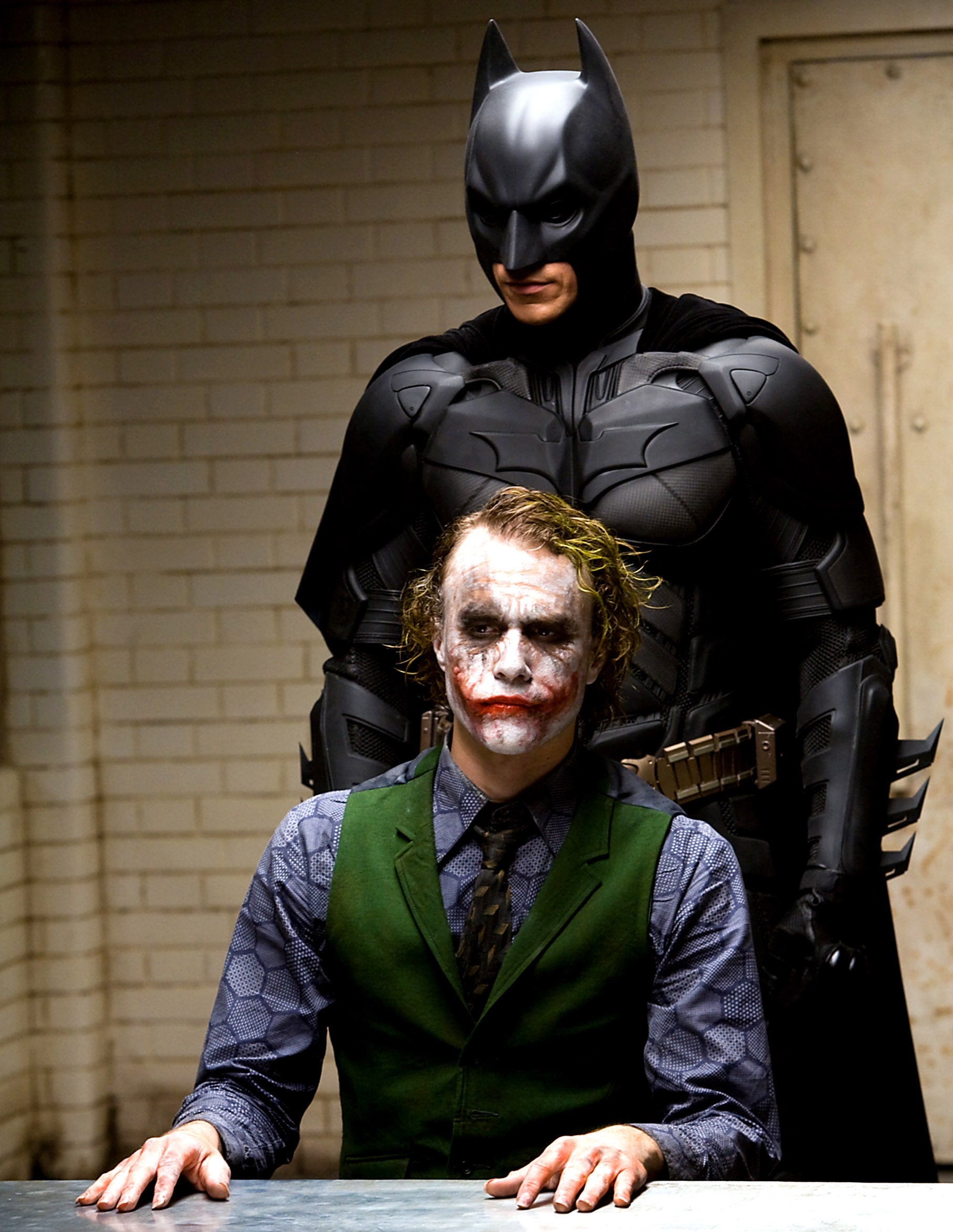 Character successful a superhero costume stands down different quality with messy constitution and a vest, seated astatine a array successful an interrogation room