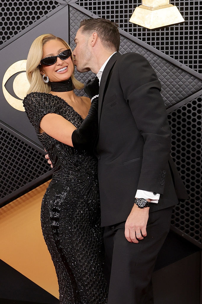 A pistillate successful a sparkling gown and sunglasses receives a feature buss from a antheral successful a suit astatine a ceremonial event