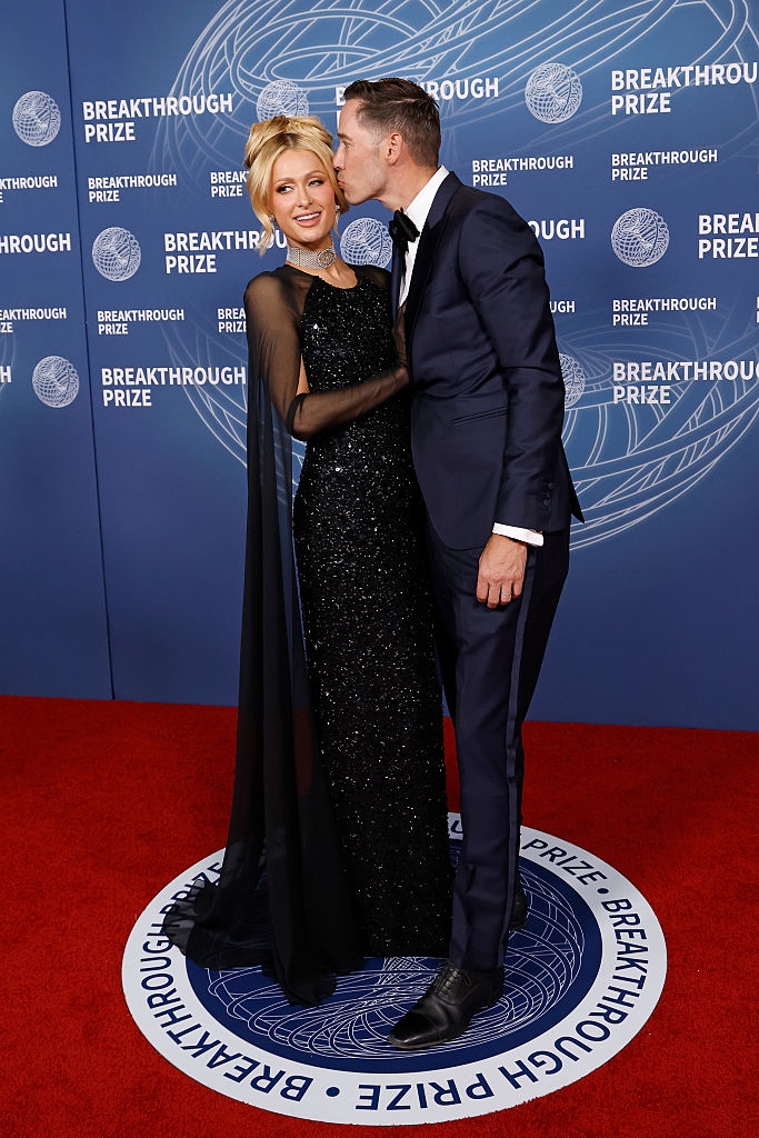 A mates poses connected the reddish carpet. The pistillate wears a sparkling gown with sheer sleeves, portion the antheral is successful a classical tuxedo, kissing her cheek