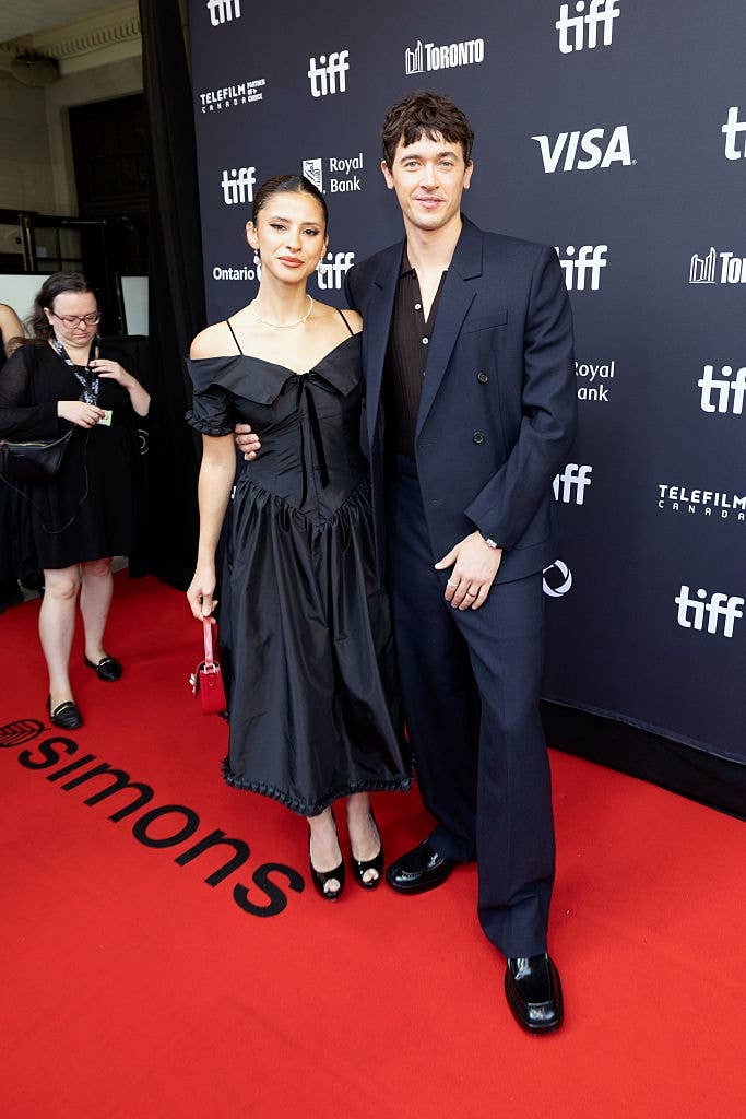 Two individuals airs  connected  a reddish  carpet astatine  a movie  event. One wears a achromatic  off-the-shoulder dress, the different   a achromatic  suit   with a acheronian  shirt. Logos successful  background