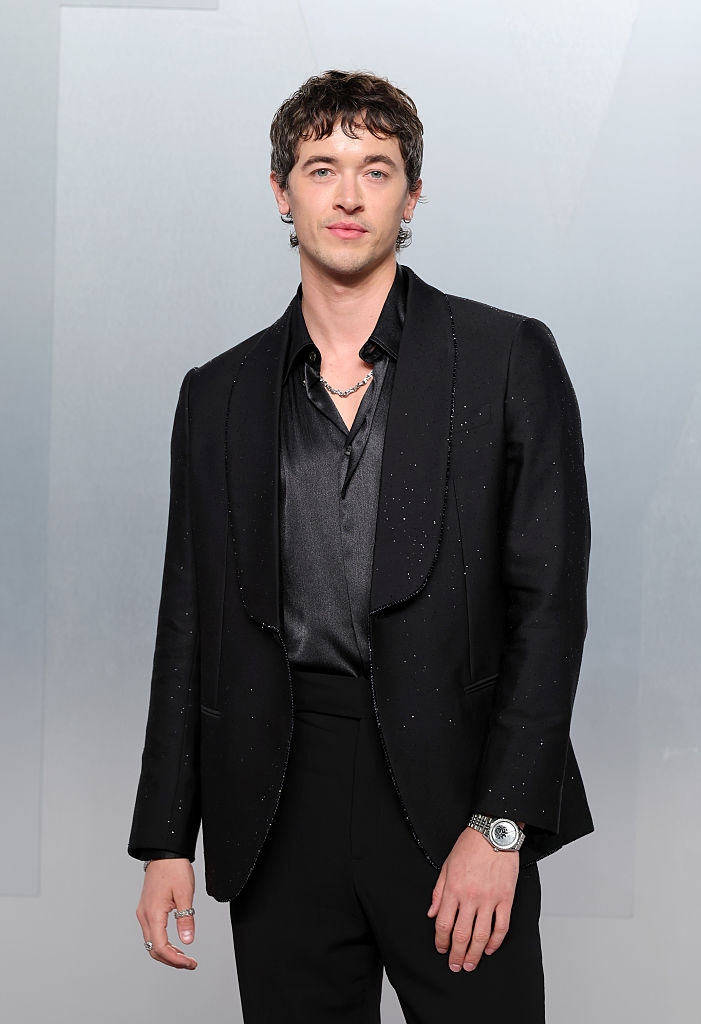 Person successful  a achromatic  suit   and shirt, with a subtle sparkle, wearing a necklace and watch, posing connected  what appears to beryllium  a ceremonial  oregon  reddish  carpet event
