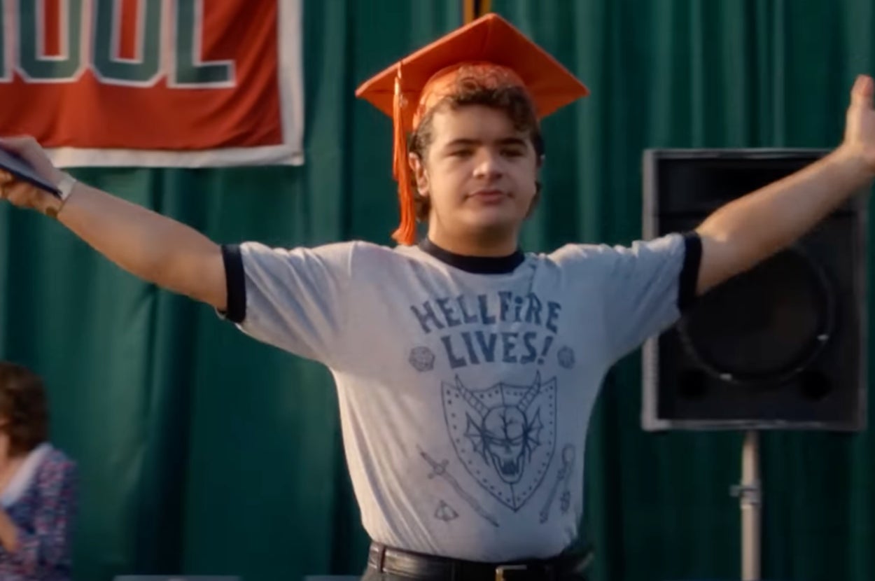 Person wearing a graduation cap and "Hellfire Lives!" T-shirt spreads arms wide, standing confidently in front of a school banner