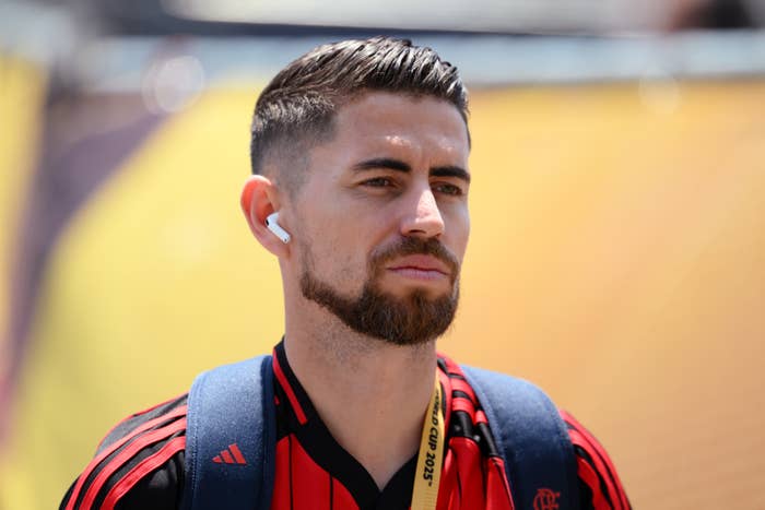 Jorginho with abbreviated  hairsbreadth  and a beard, wearing earbuds, a sports jersey, and a backpack, appears focused portion    outdoors