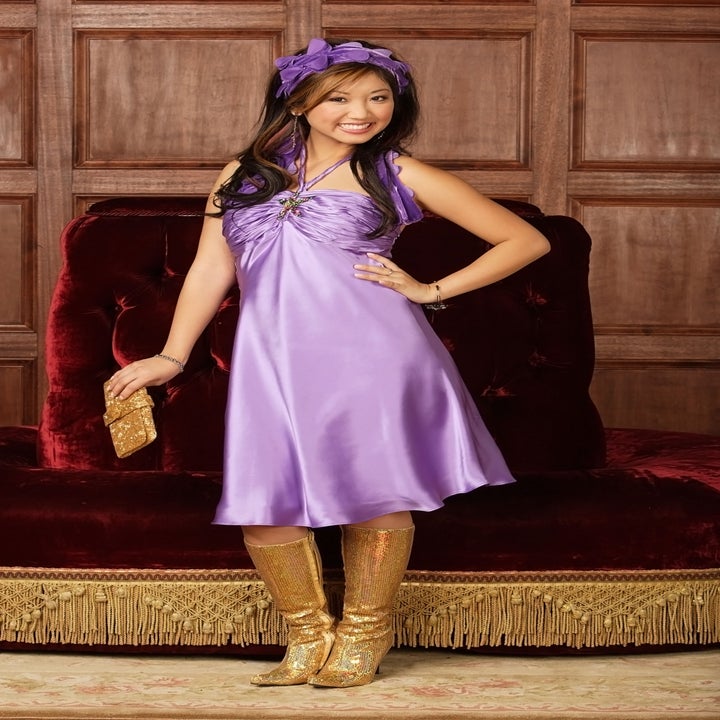 Person in a stylish, sleeveless satin dress and gold boots, holding a matching purse, poses confidently on a velvet backdrop