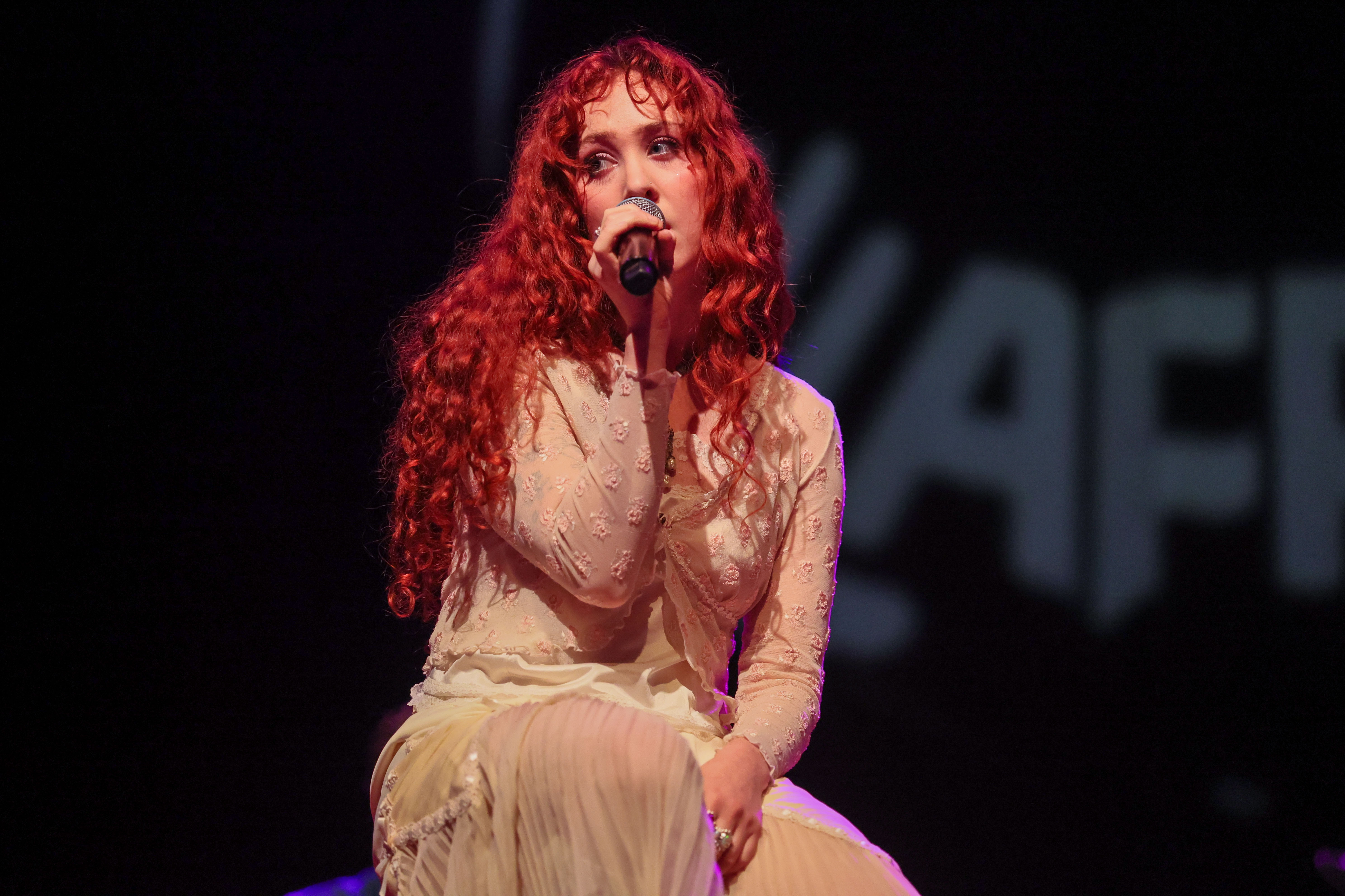 Person with agelong curly hairsbreadth successful an elegant, sheer outfit performs connected stage, holding a microphone