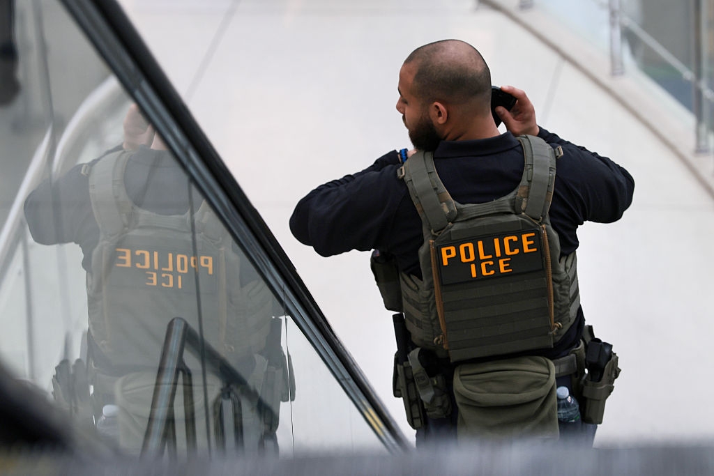 An ICE officer, wearing a tactical vest labeled "POLICE ICE," stands connected an escalator, adjusting his earpiece