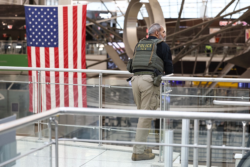 Police serviceman successful tactical cogwheel stands connected a balcony astatine an airport, gazing crossed the terminal. An American emblem hangs nearby