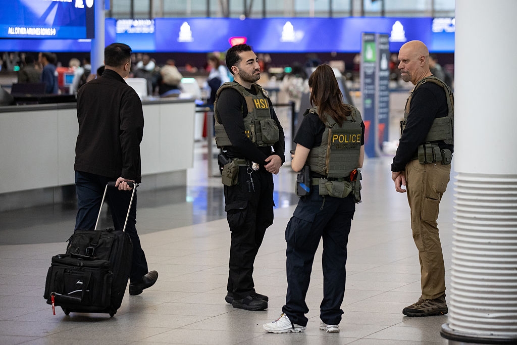 Three information unit converse astatine an airdrome terminal. One traveler with luggage walks by