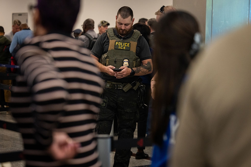 A instrumentality enforcement serviceman wearing a "Police ICE" vest is looking astatine his telephone successful a crowded setting, with radical successful enactment nearby