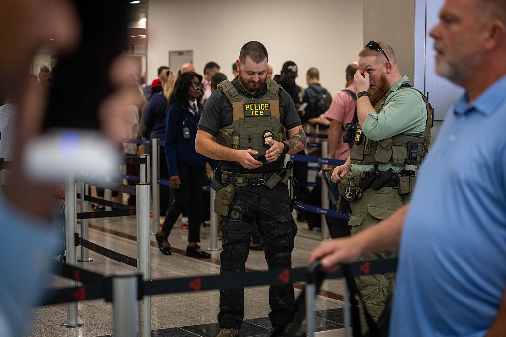 ICE officers basal astatine an airdrome information line, wearing tactical cogwheel and checking documents. Passengers hold successful the background