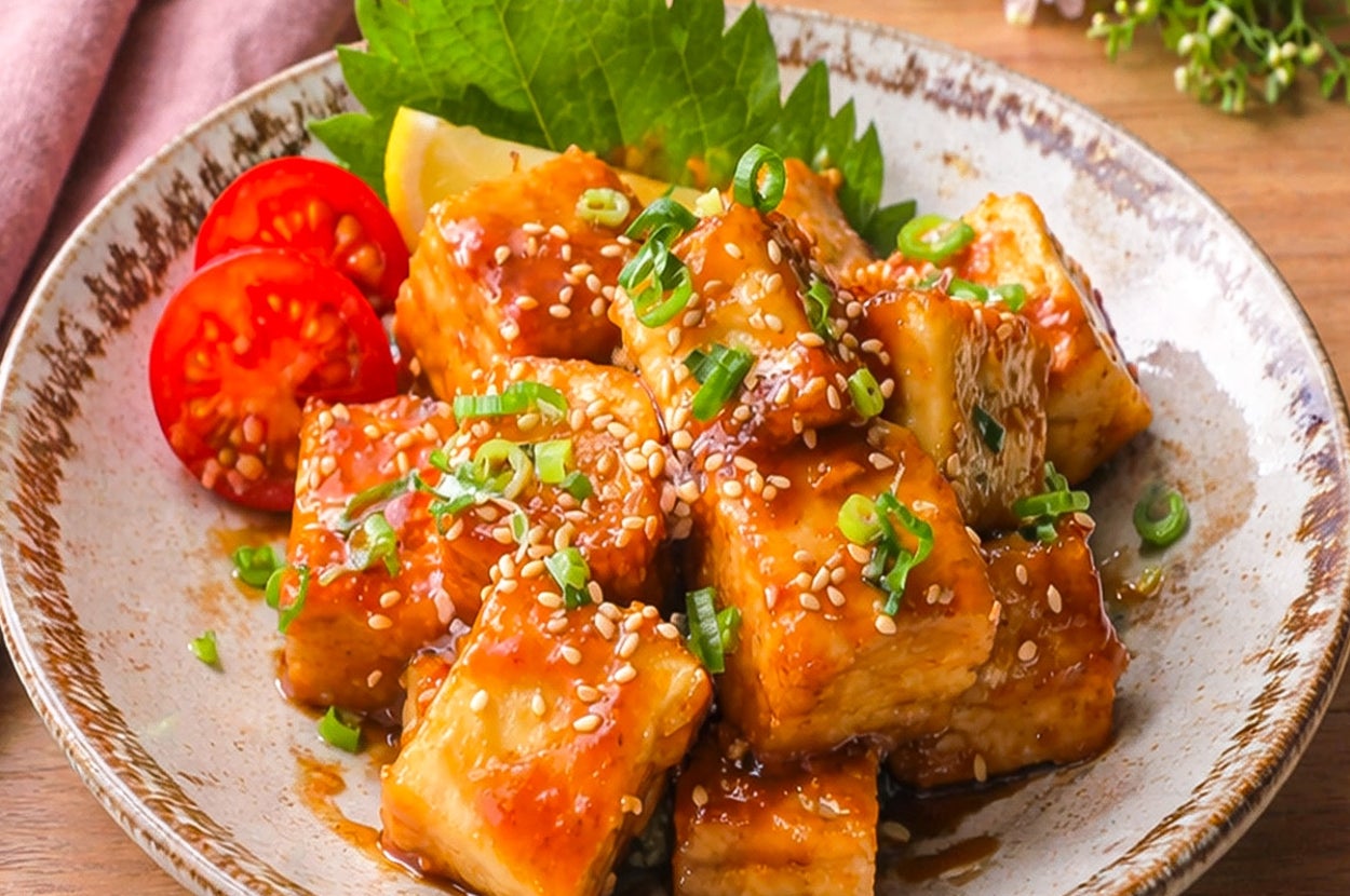 焼き豆腐の揚げ物が白い皿に盛られ、ゴマとネギがトッピングされている。皿の背景に花と布製ナプキンがある。