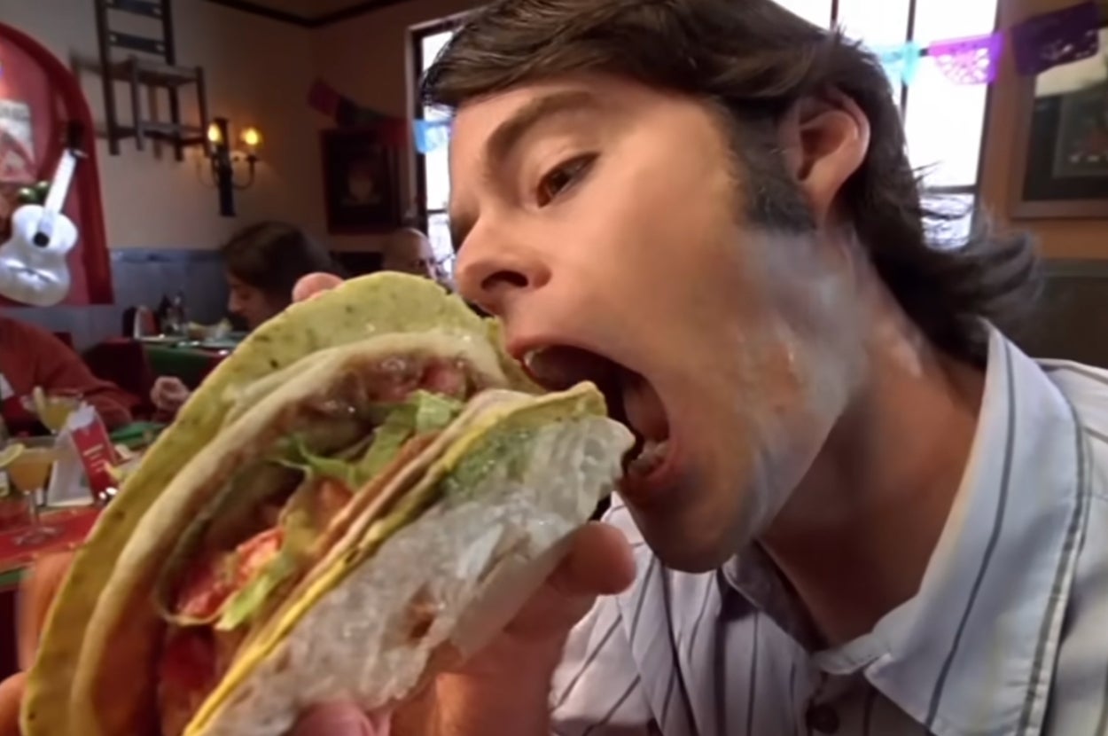 A person takes a big bite of a large, stuffed taco at a restaurant. Diners and festive decorations are seen in the background