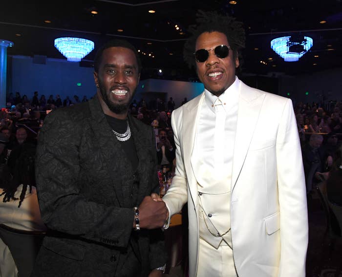 Two individuals smiling and shaking hands astatine  a ceremonial  event. One wears a textured suit; the different   is successful  a sleek achromatic  suit   with sunglasses