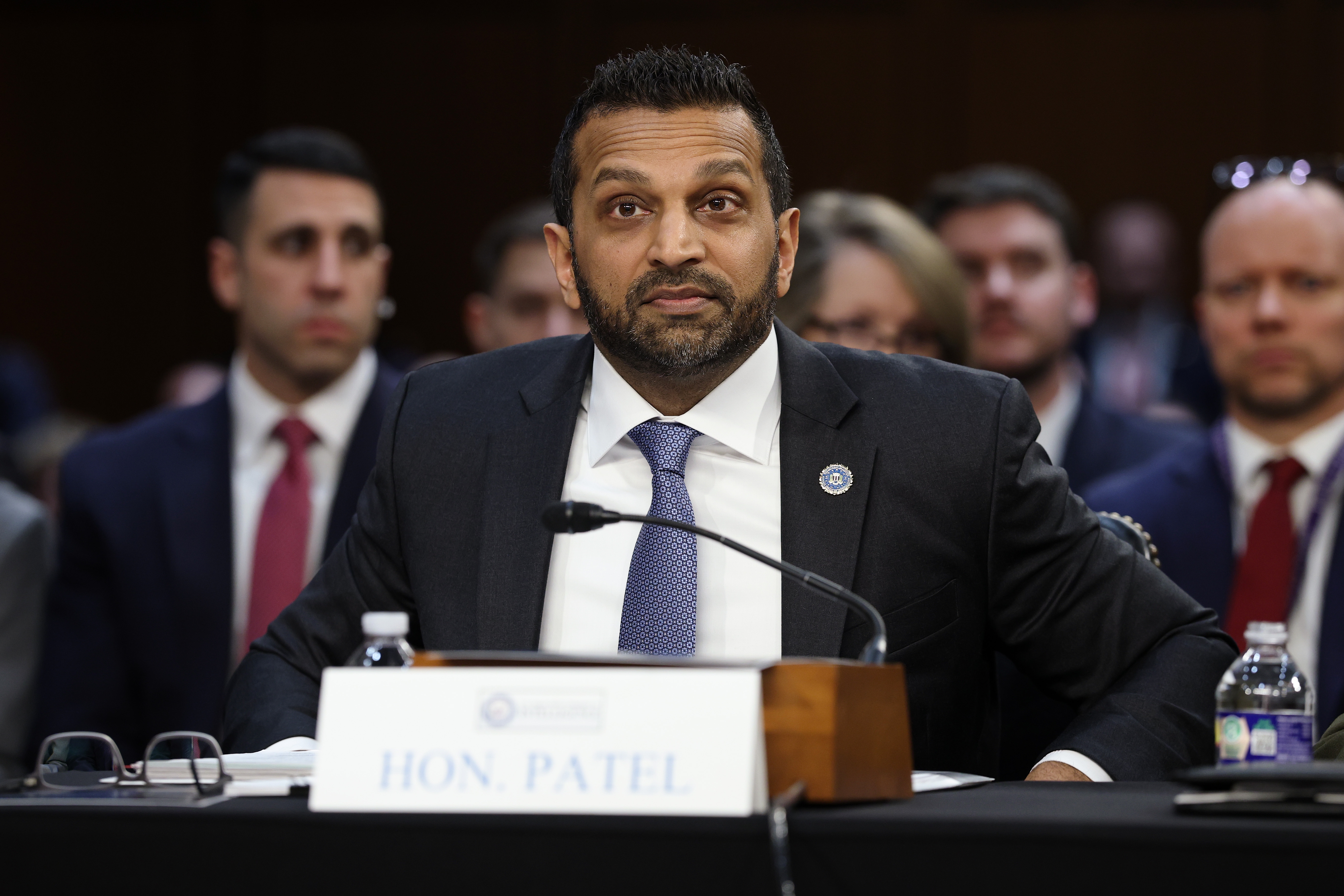 A seated idiosyncratic  successful  a suit   listens intently during a ceremonial  hearing, with a nameplate that reads "Hon. Patel" successful  beforehand   of them