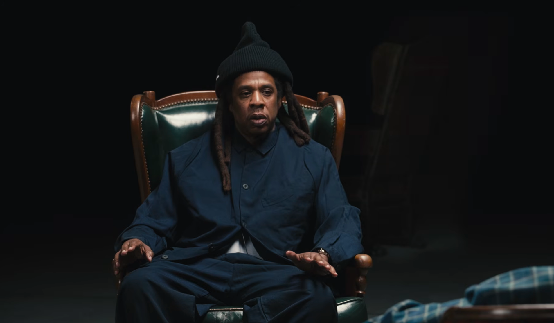 A idiosyncratic   with agelong  hairsbreadth  sits successful  a leather chair, wearing a acheronian  beanie and loose-fitting clothing