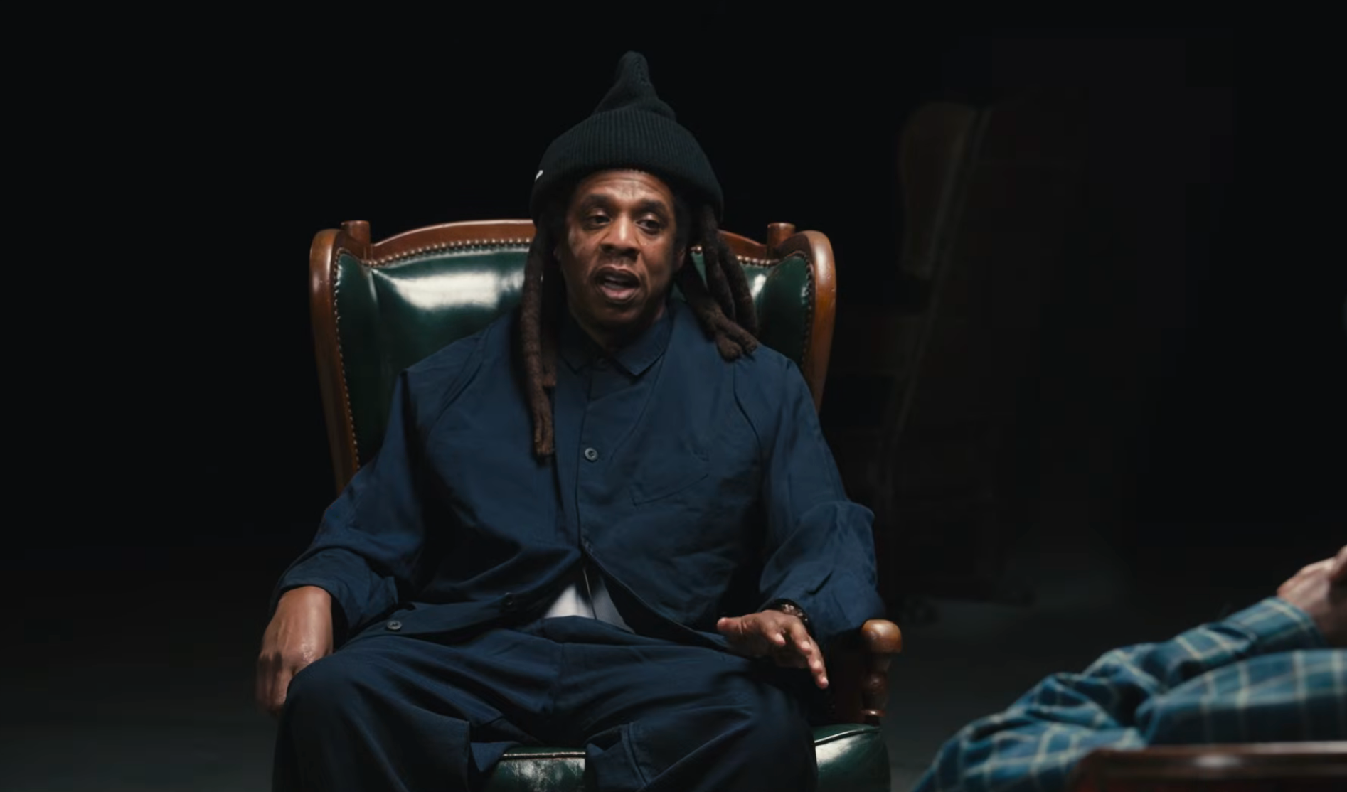 Person successful  a acheronian  outfit and beanie sits successful  a ample  chair, gesturing portion    speaking